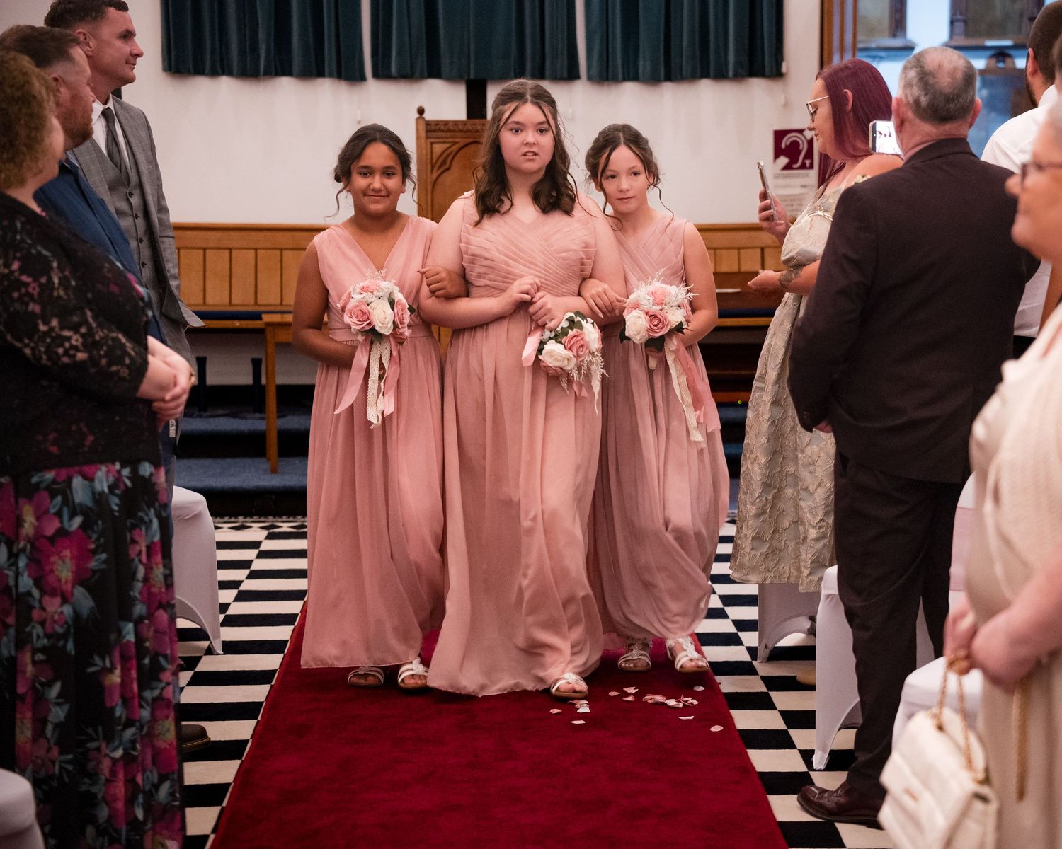 Three bridesmaids in pink gowns walk down a red carpet aisle holding bouquets during a wedding ceremony.