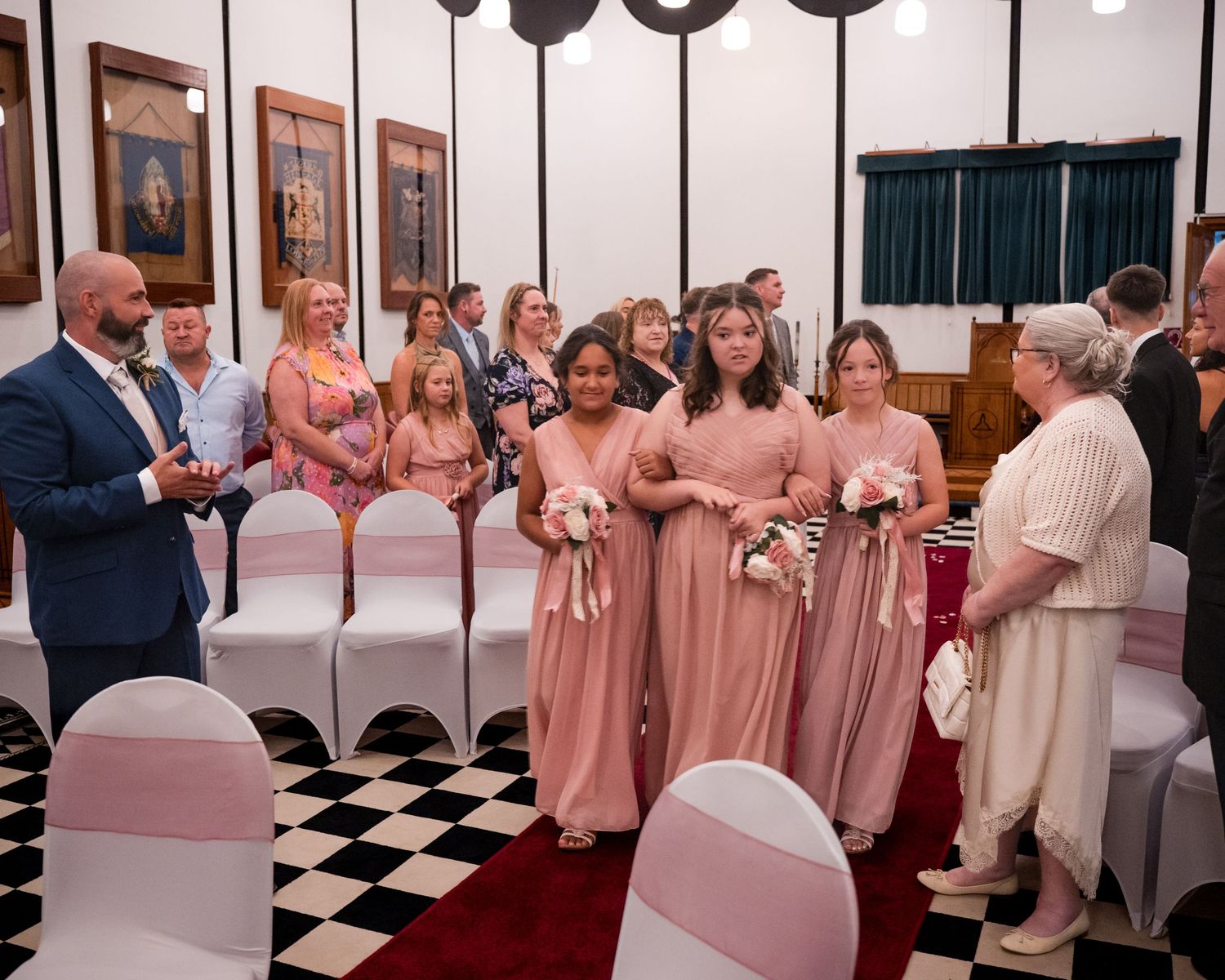 Bridesmaids in blush pink gowns walk down the aisle at a wedding ceremony in a decorated venue.