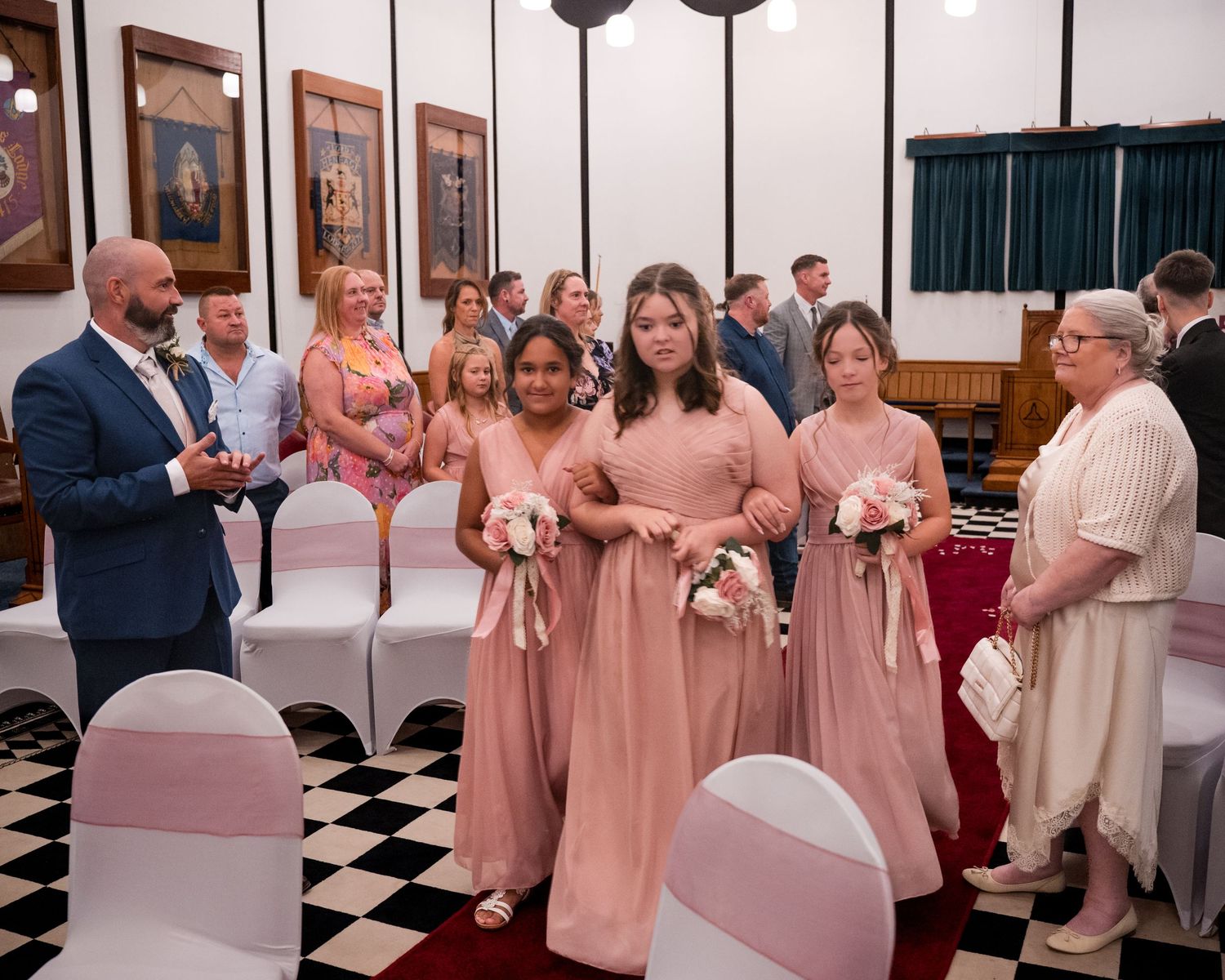 Bridesmaids in pink dresses walk down the aisle at a wedding ceremony in a decorated hall.