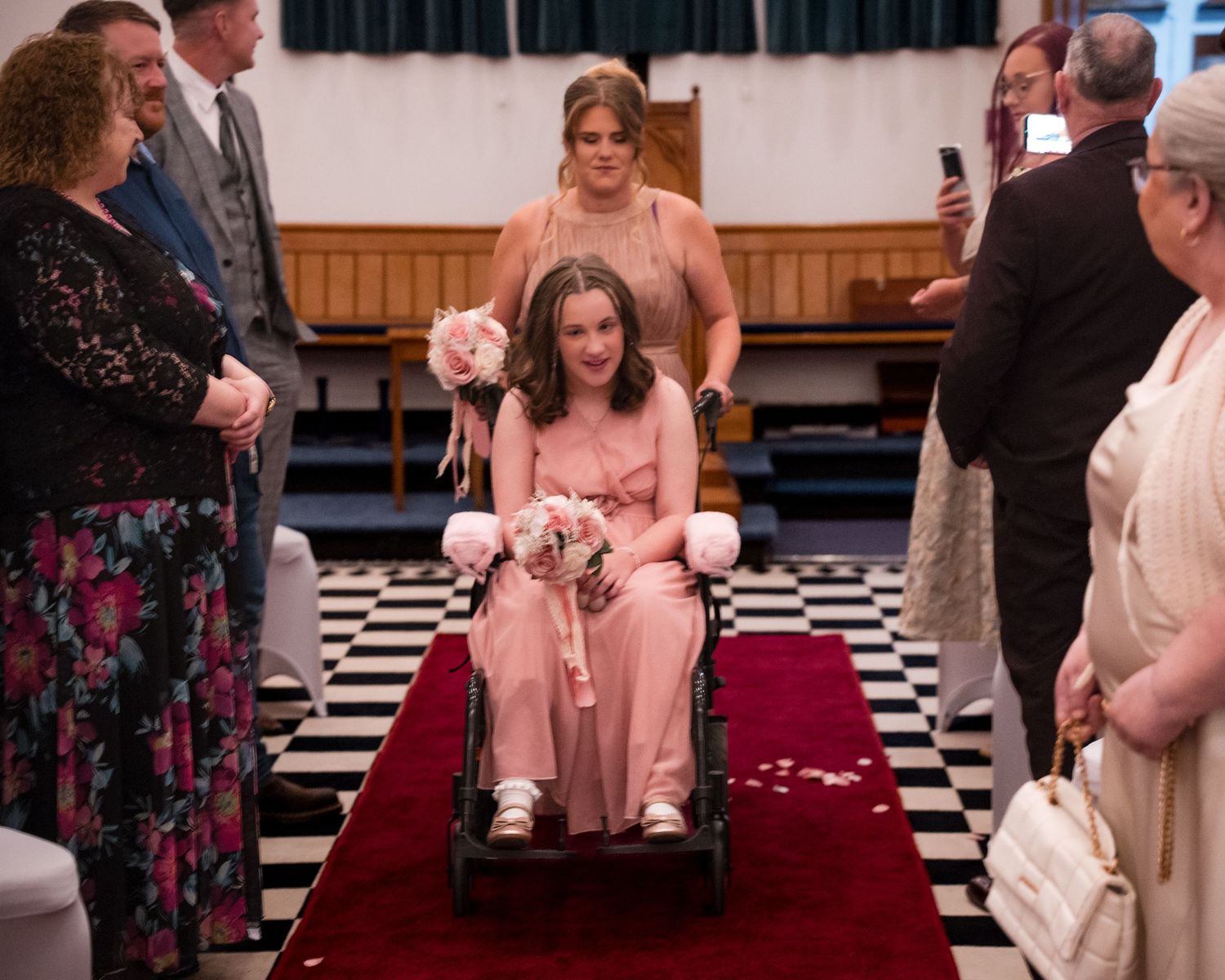 Bridesmaid in pink dress using wheelchair rolls down red carpet aisle in church wedding ceremony.