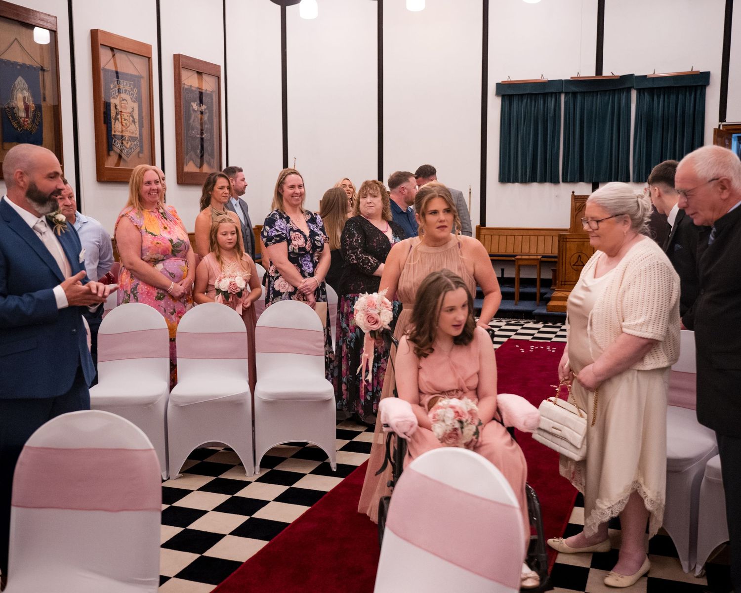 Wedding ceremony scene with guests seated on white chairs in a checkered floor hall, flower girls walking the aisle.