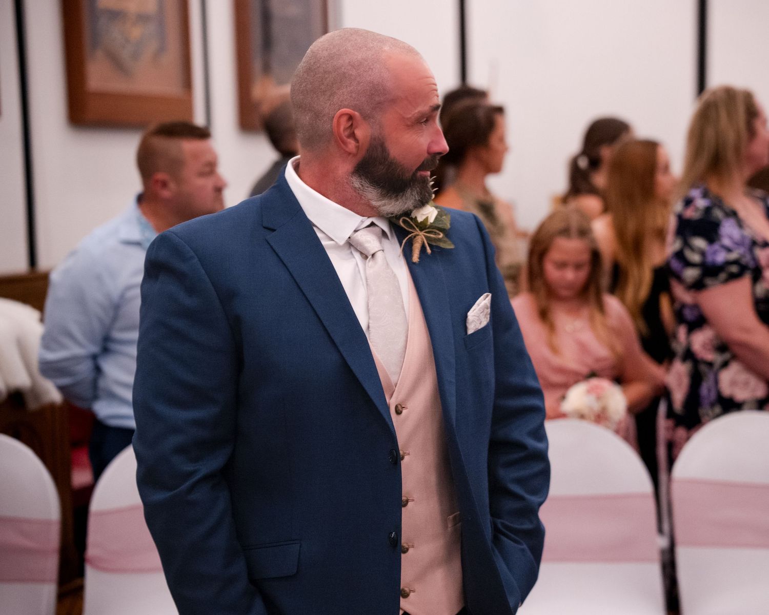 Bald man with beard in blue suit and bow tie stands at wedding ceremony with guests seated in background.