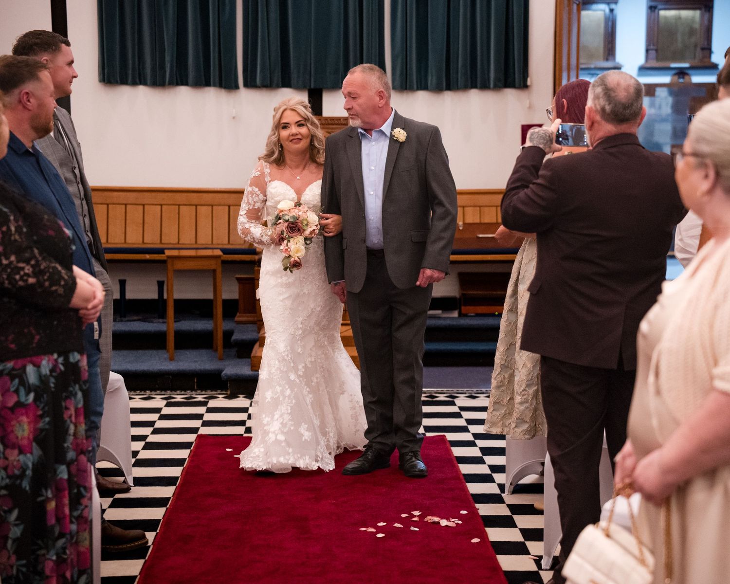 Bride in white lace gown walks down red carpet aisle with escort at elegant wedding ceremony.