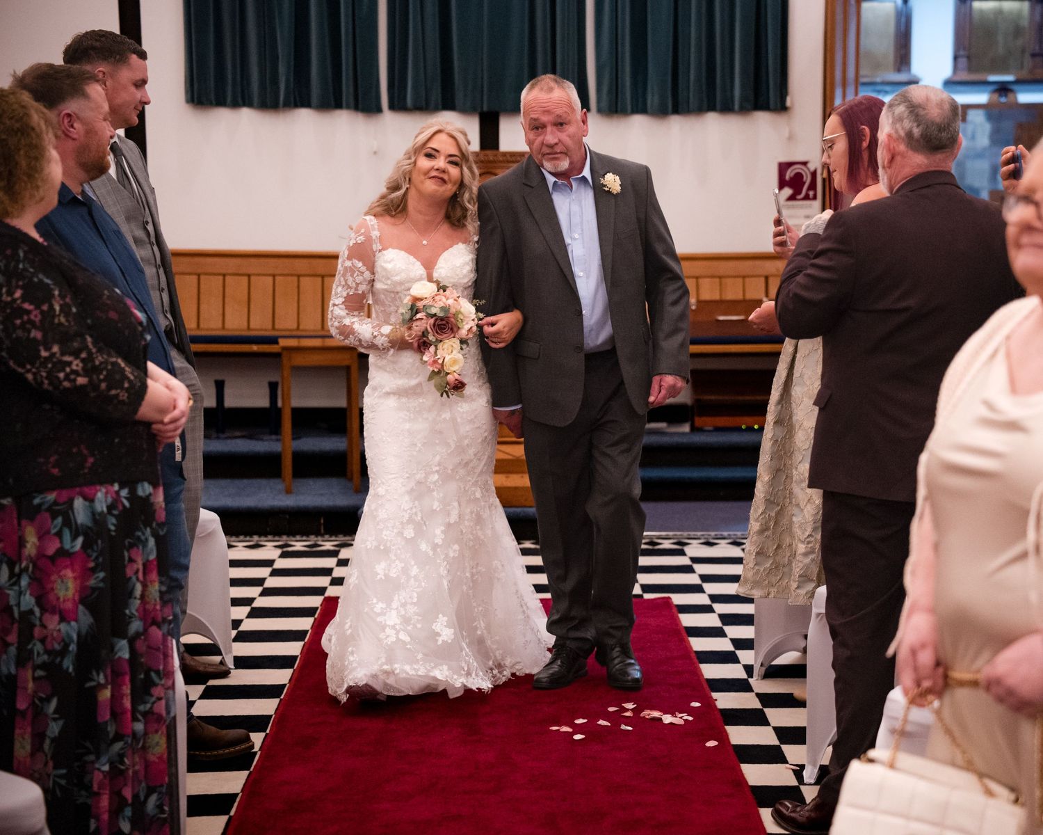 Bride in lace wedding gown walks down red carpet aisle with escort at indoor wedding ceremony.