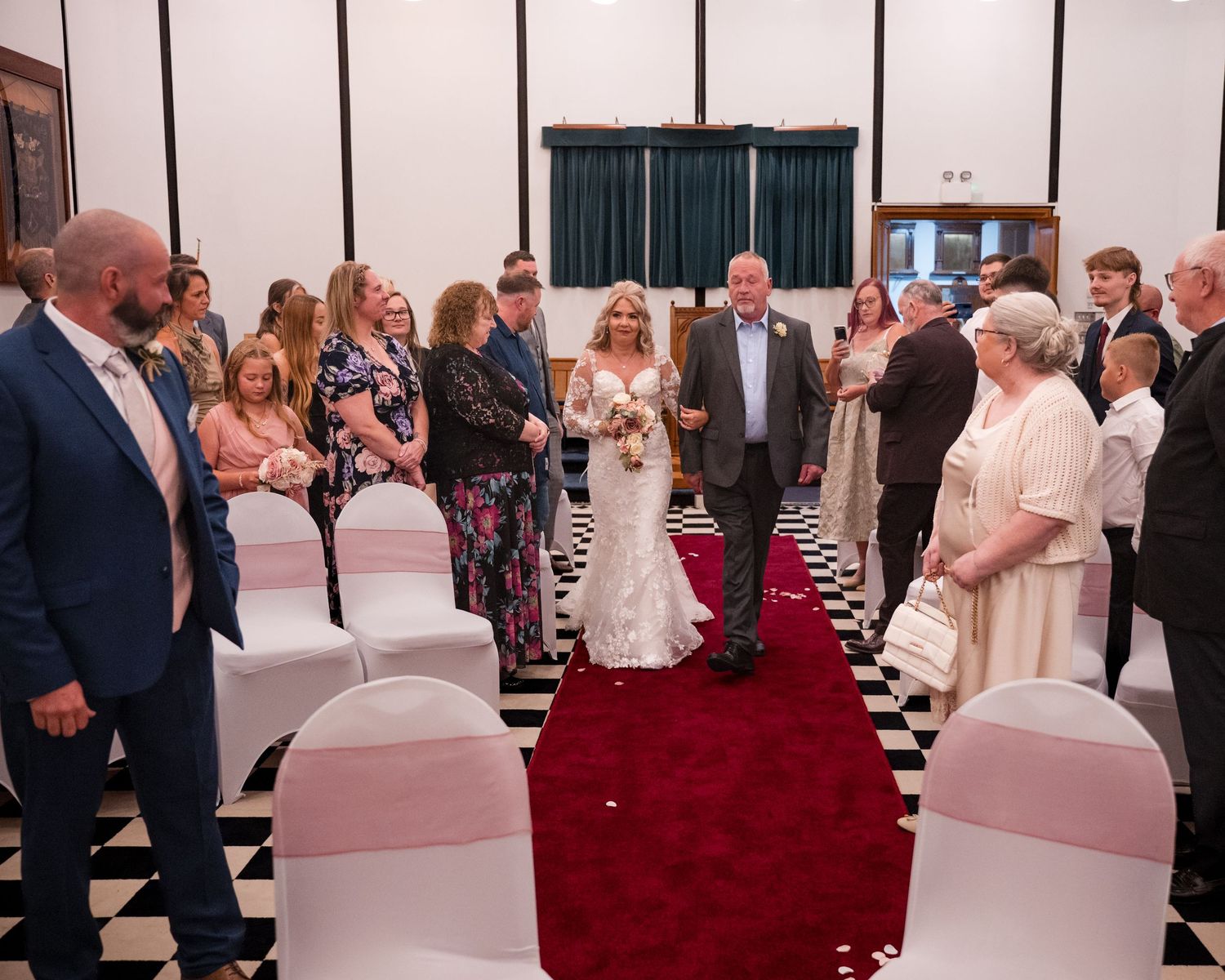 Bride walks down the aisle in a white gown on a red carpet, surrounded by guests in a decorated wedding venue.