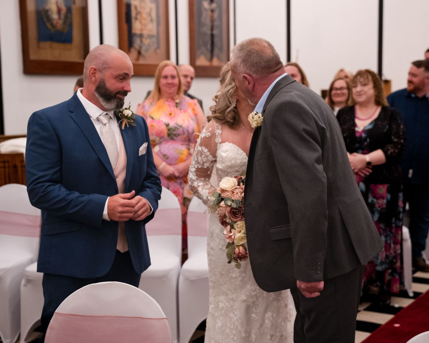 Groom in navy suit greets bride holding bouquet at wedding ceremony altar with guests seated in background.