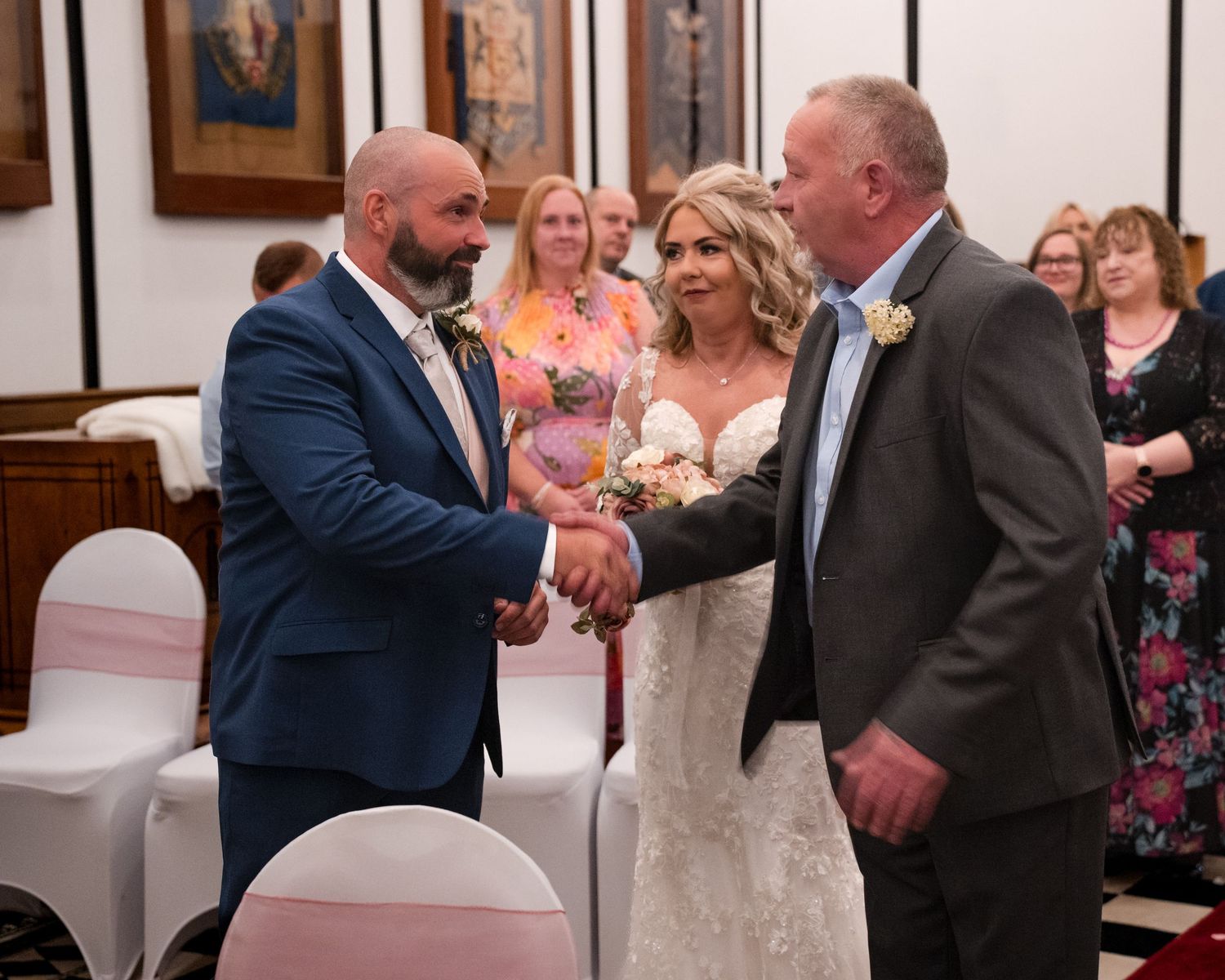 Bride in lace gown stands between two men shaking hands at a wedding ceremony in a decorated venue.