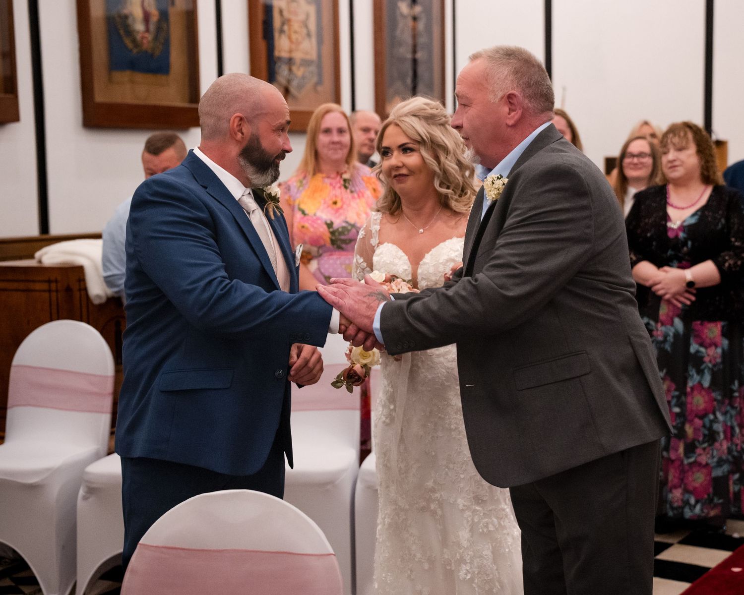 Bride in lace gown stands between two men in suits shaking hands at a wedding ceremony in a decorated venue.