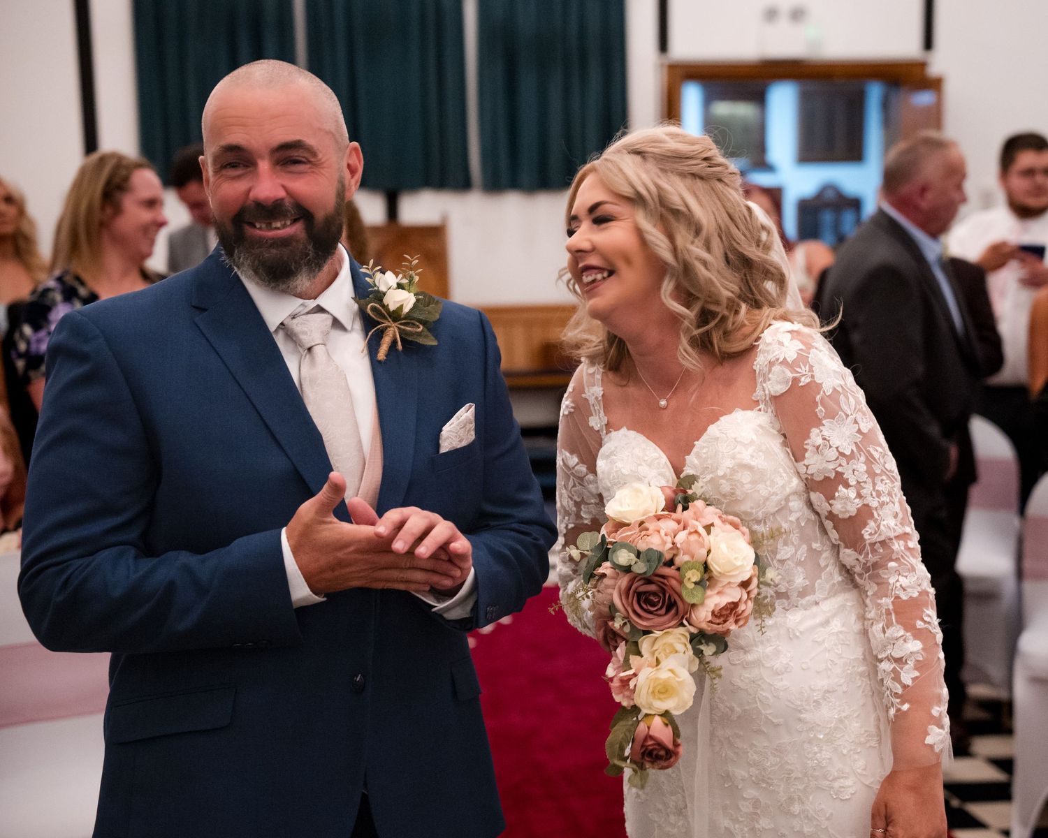 Smiling bride in lace gown holding bouquet walks with groom in navy suit during wedding ceremony.