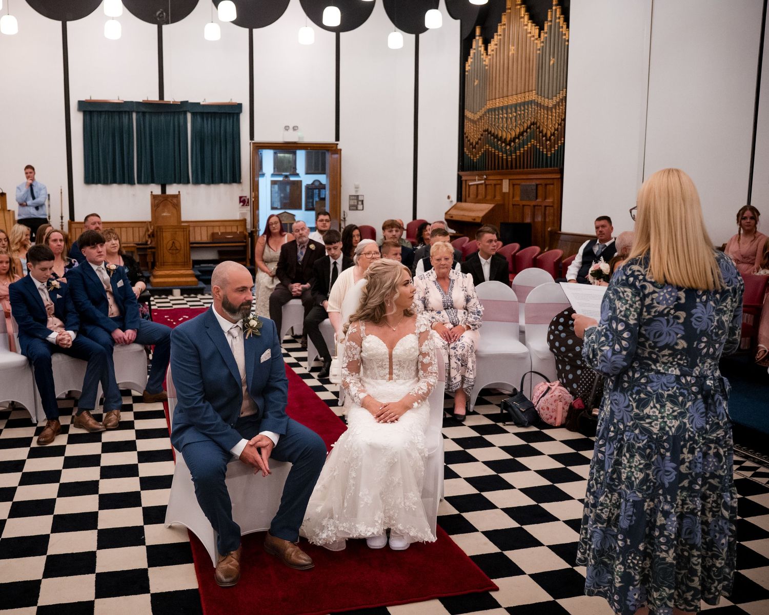 Bride and groom seated during wedding ceremony on checkered floor, guests watching as photographer captures the moment.