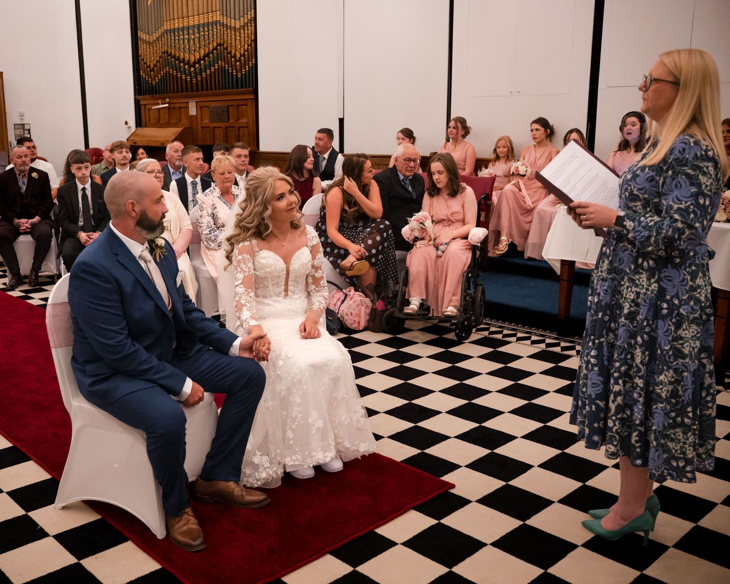 Bride and groom seated during wedding ceremony on checkered floor, officiant reading vows, guests watching in decorated venue.