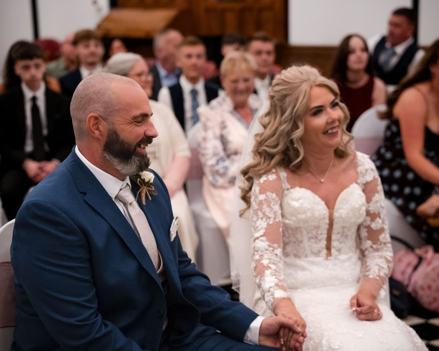 Bride and groom smiling during wedding ceremony, bride in lace dress, groom in blue suit, guests seated in background.
