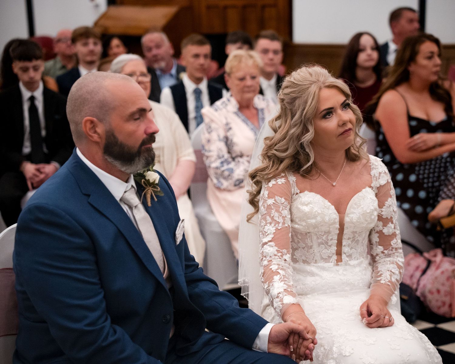 Bride and groom sitting together at wedding ceremony altar, bride in lace dress, groom in navy suit, guests seated behind.
