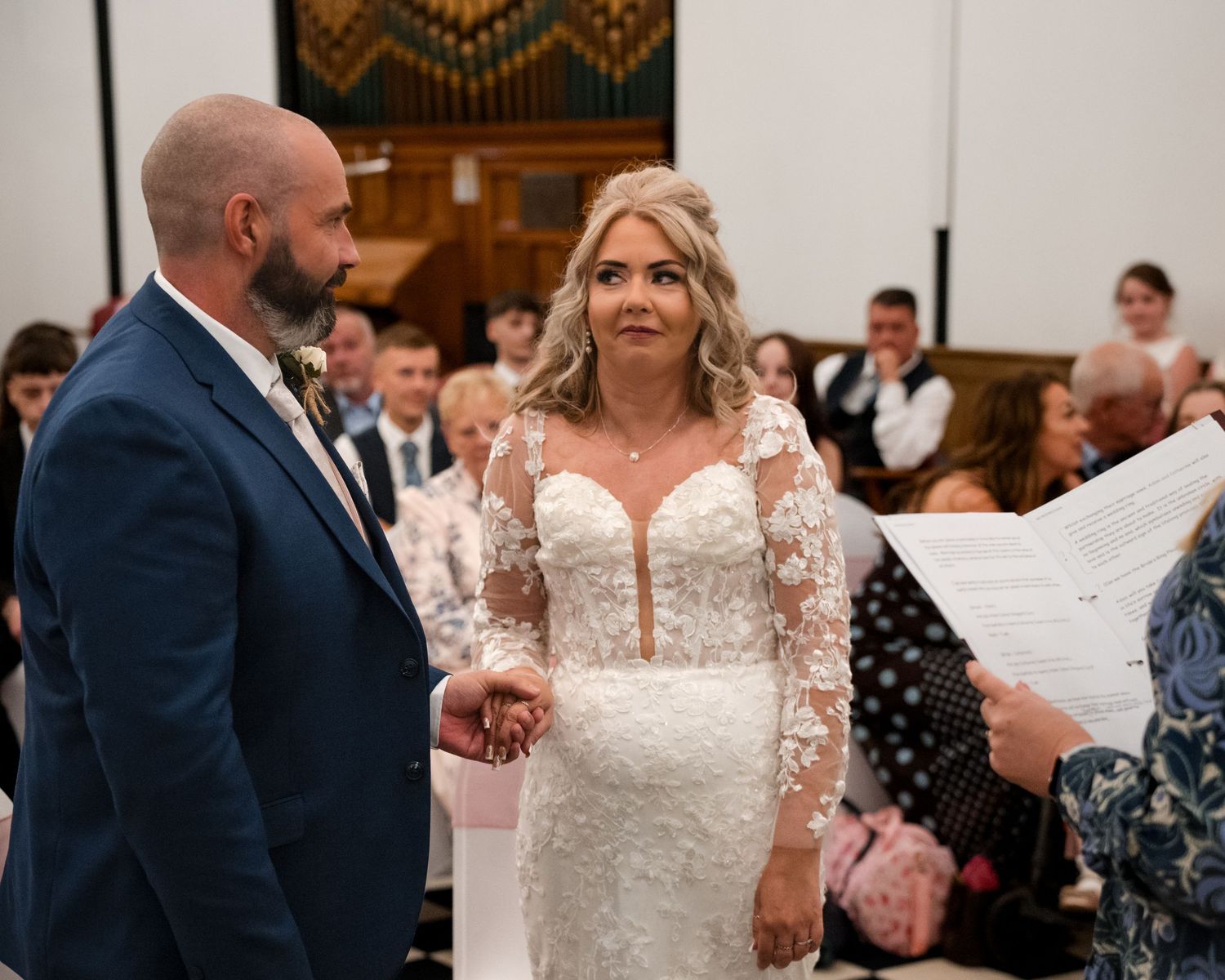 Bride in lace wedding dress holds hands with groom in blue suit during church wedding ceremony.