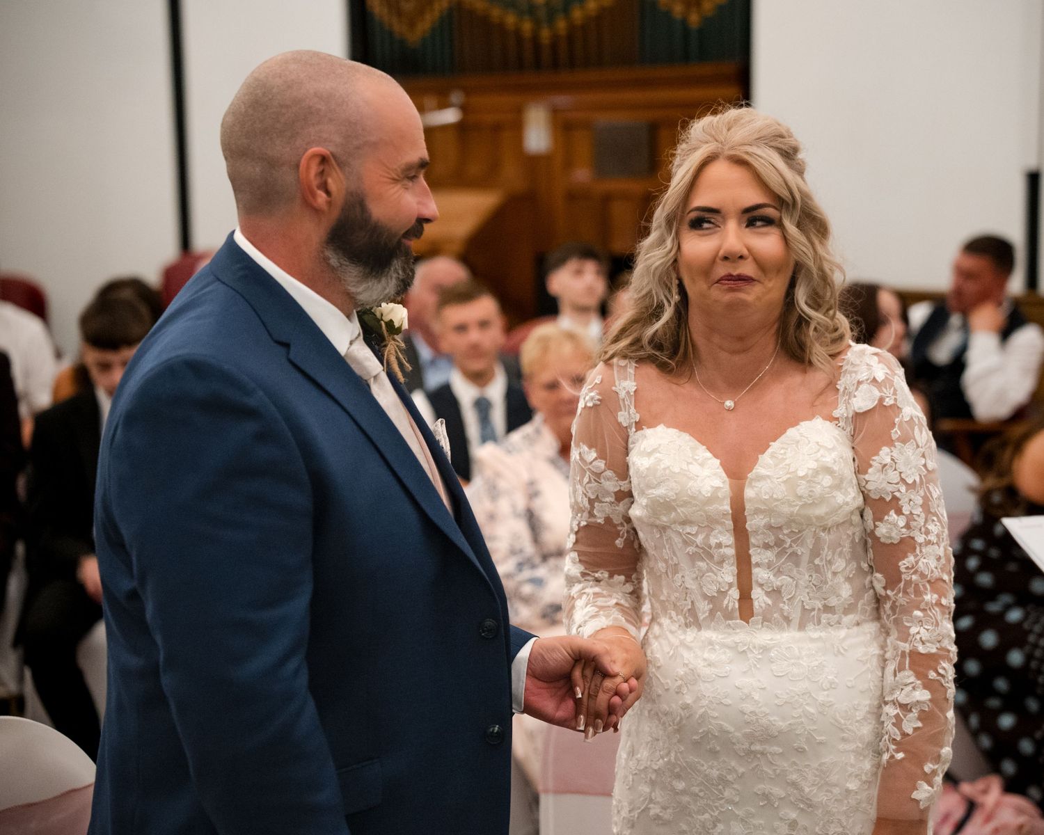 A bride and groom hold hands during their wedding ceremony in a church, bride wearing lace gown, groom in blue suit.