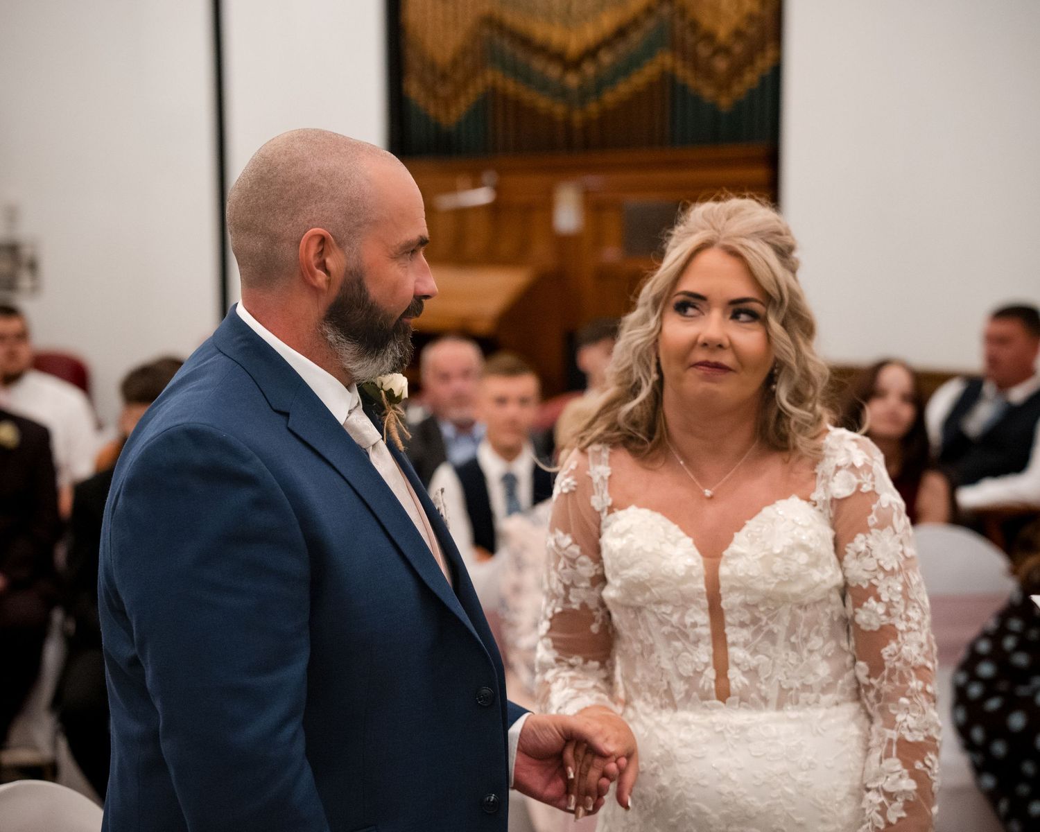 Bride and groom holding hands during wedding ceremony in church, bride wearing lace long-sleeve gown, groom in navy suit.