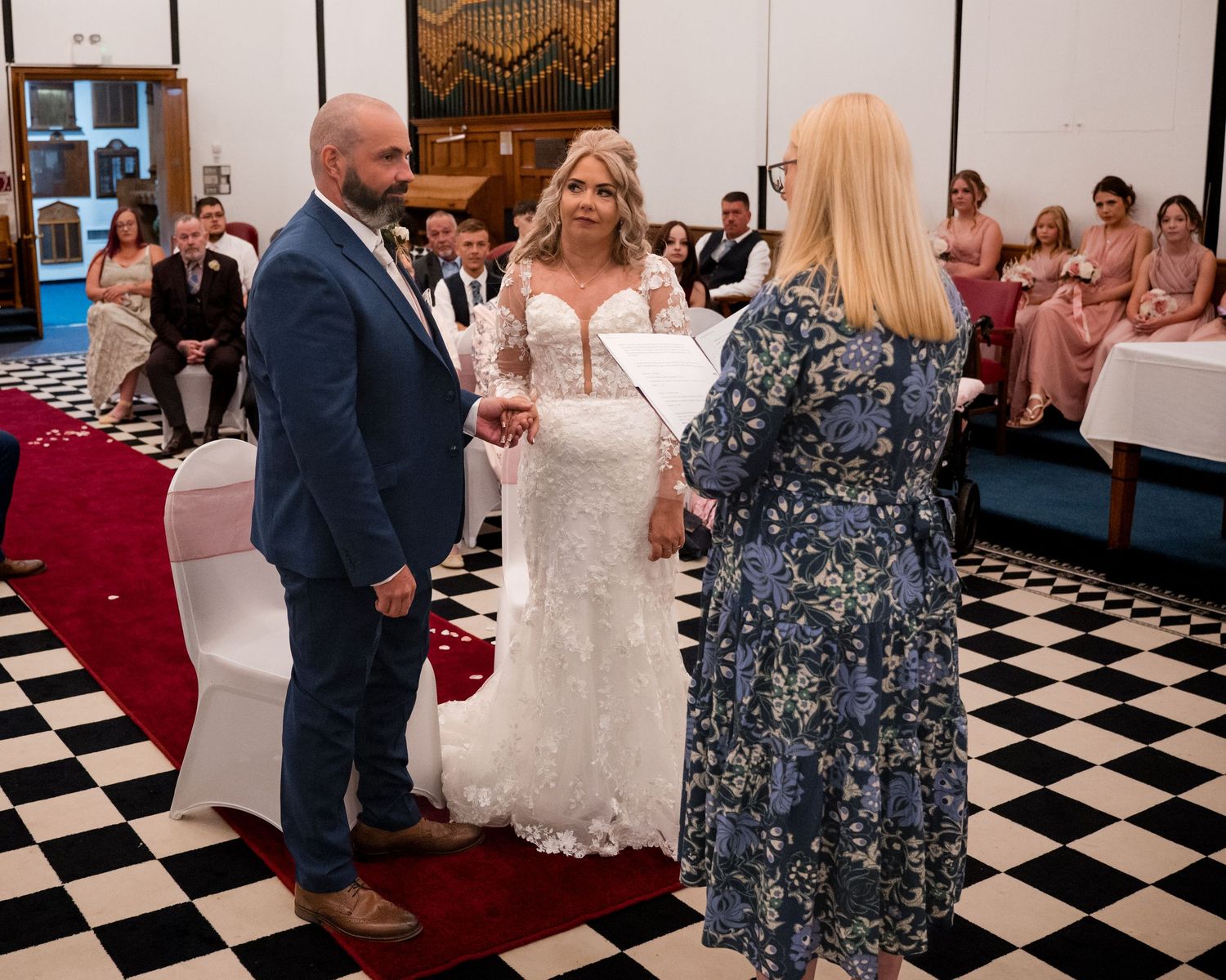 Bride and groom exchanging vows with officiant on checkered floor, wedding guests seated in background.