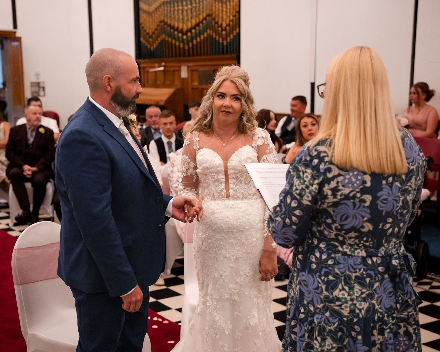 Bride and groom exchange vows at wedding ceremony, officiant holding papers, guests seated in decorated venue.