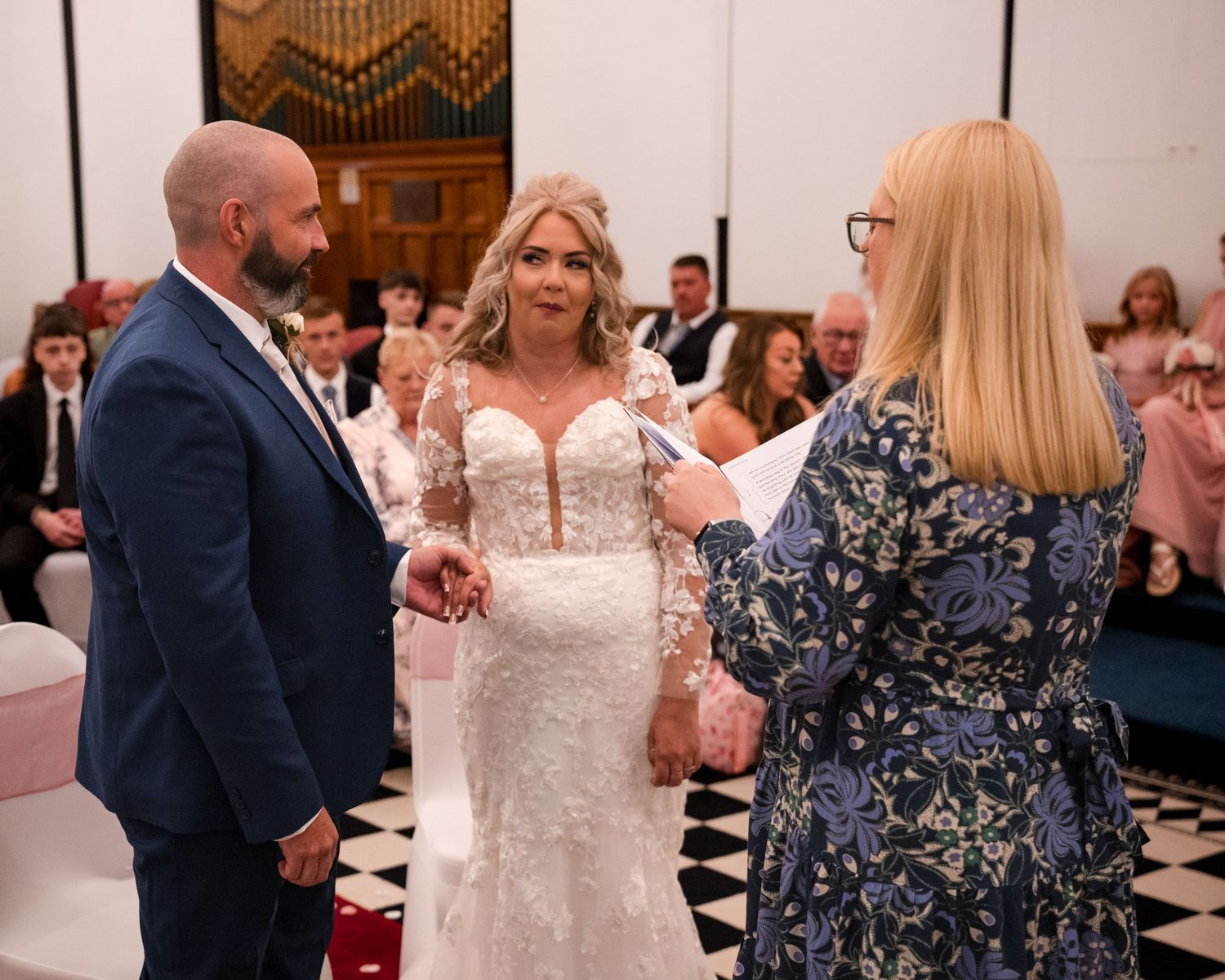 Bride and groom exchanging vows during wedding ceremony with officiant reading from paper in elegant venue.
