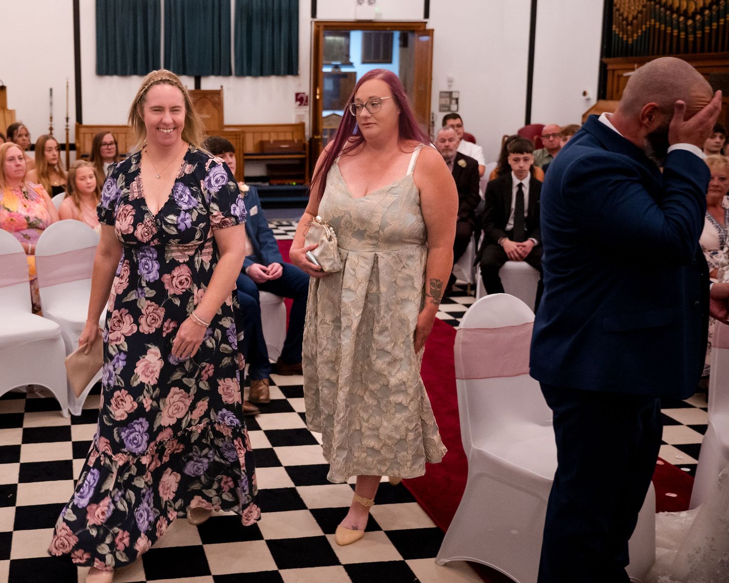 Two women in elegant dresses walk through a decorated hall during a formal wedding ceremony.
