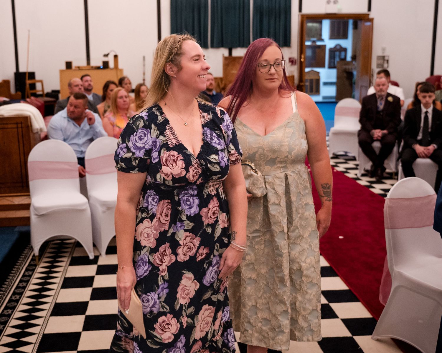 Two women walking down a church aisle at a wedding ceremony, guests seated on white chairs in background.