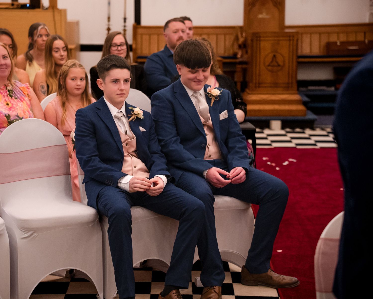 Two young men in navy suits sit at a wedding ceremony in a church, wearing boutonnieres.
