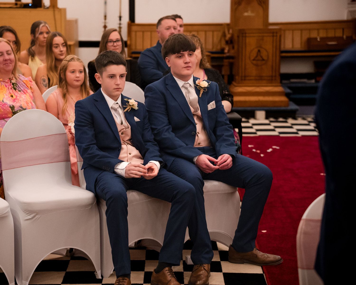 Two young men in navy suits with floral buttonholes seated at a wedding ceremony in a church.