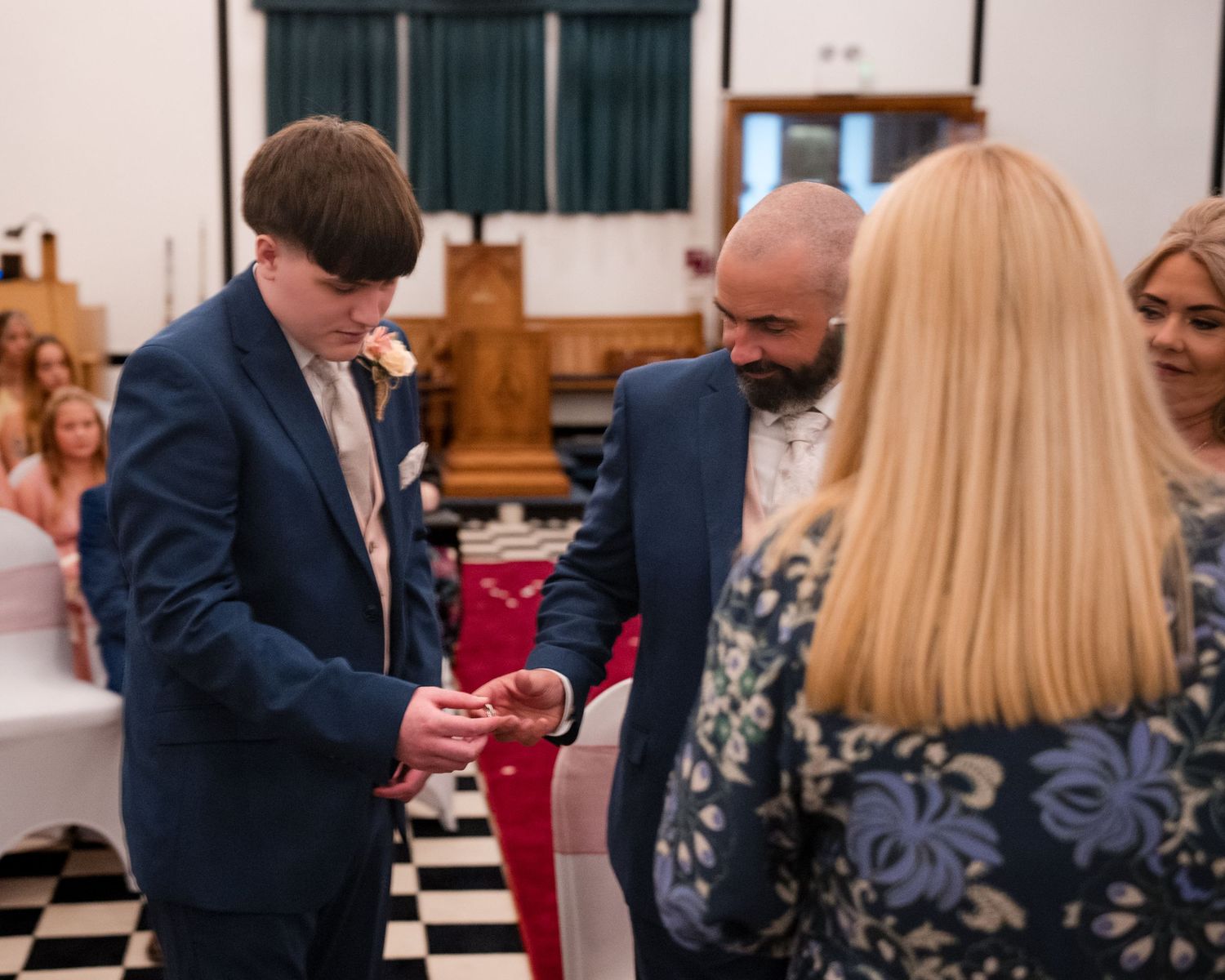 Two grooms exchange rings during a wedding ceremony in a hall with black and white checkered floors.