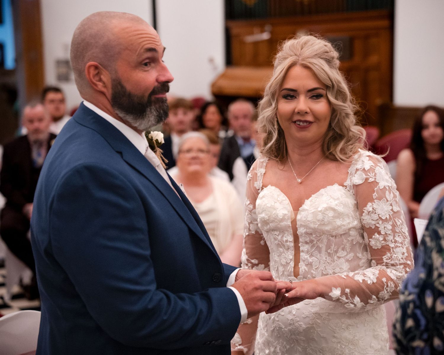 A bride and groom exchange rings during their wedding ceremony, with guests seated in the background.