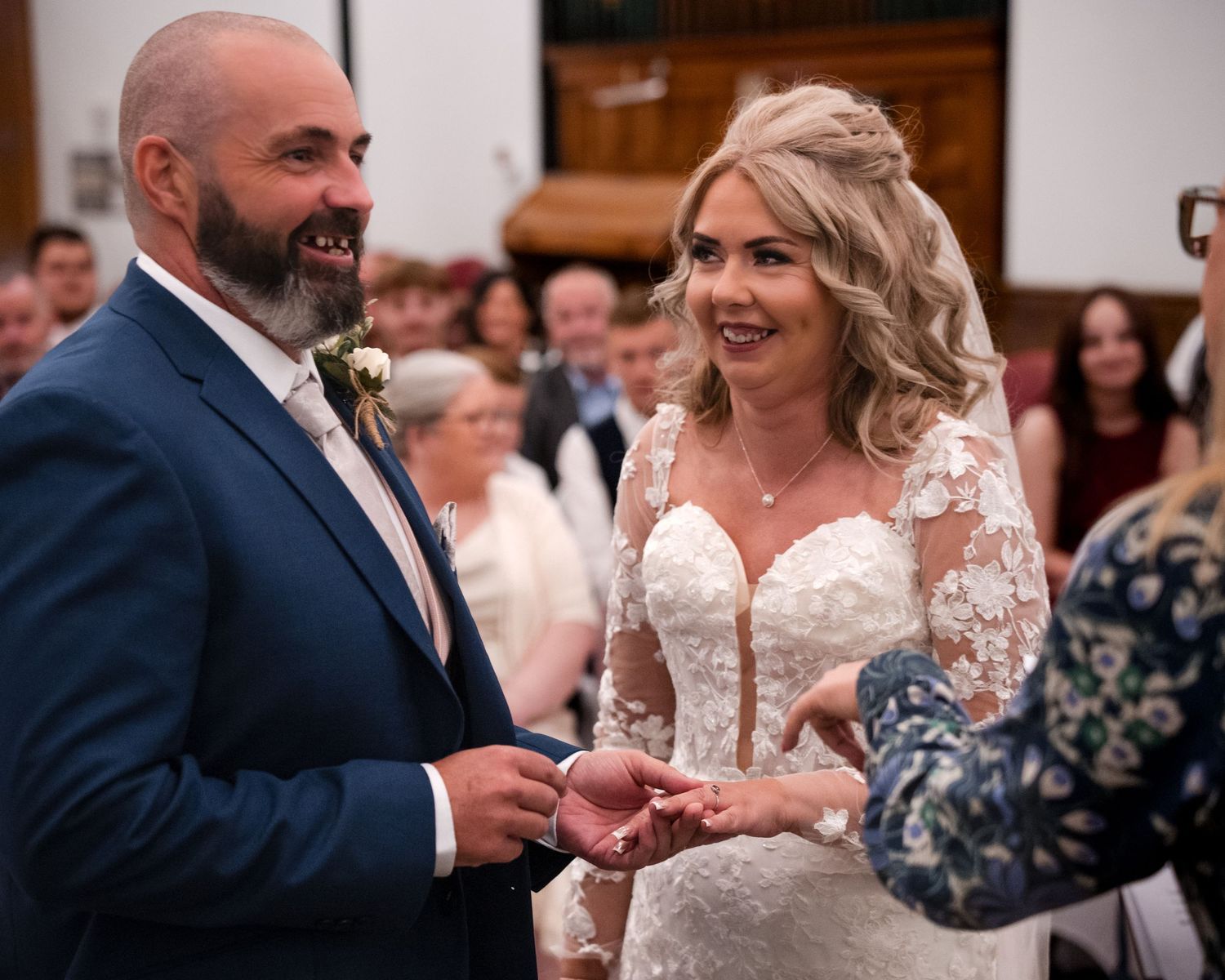 Bride and groom exchanging rings at wedding ceremony, smiling joyfully, bride in lace dress, groom in blue suit.