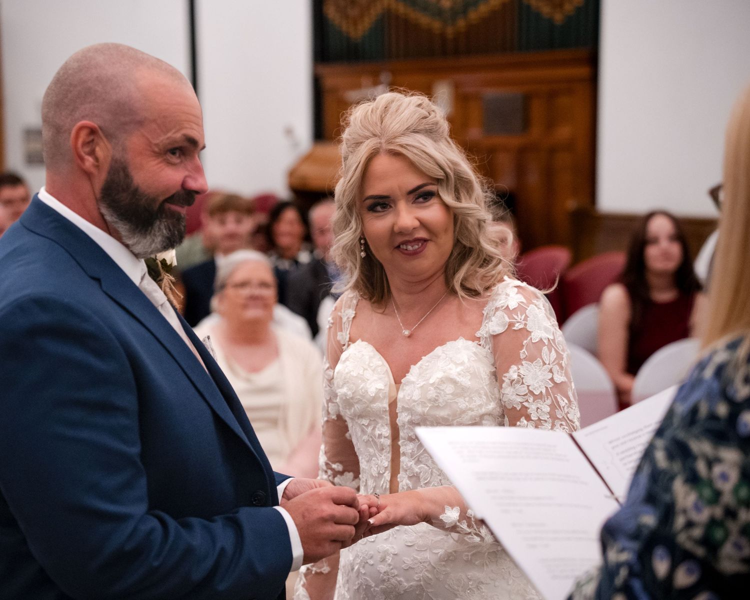 Couple exchanging wedding rings during civil ceremony, bride in lace gown smiling at groom in navy suit.