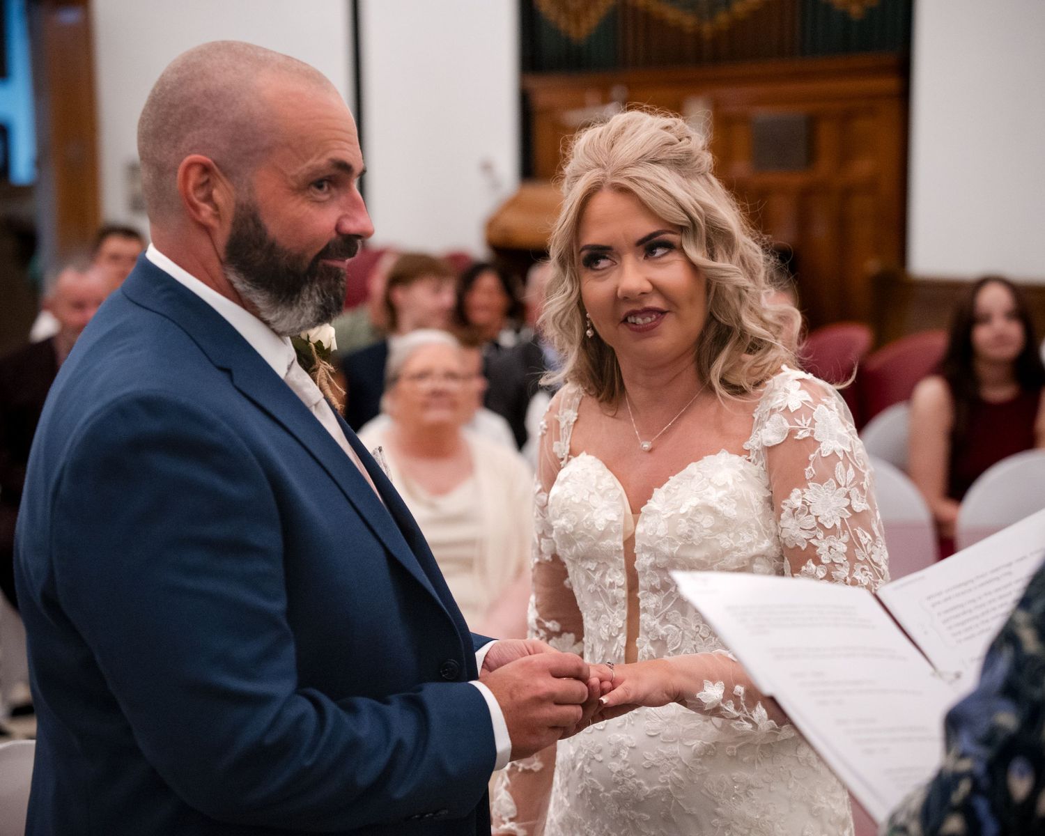 Bride and groom exchange vows at wedding ceremony, bride wearing lace gown, groom in blue suit.