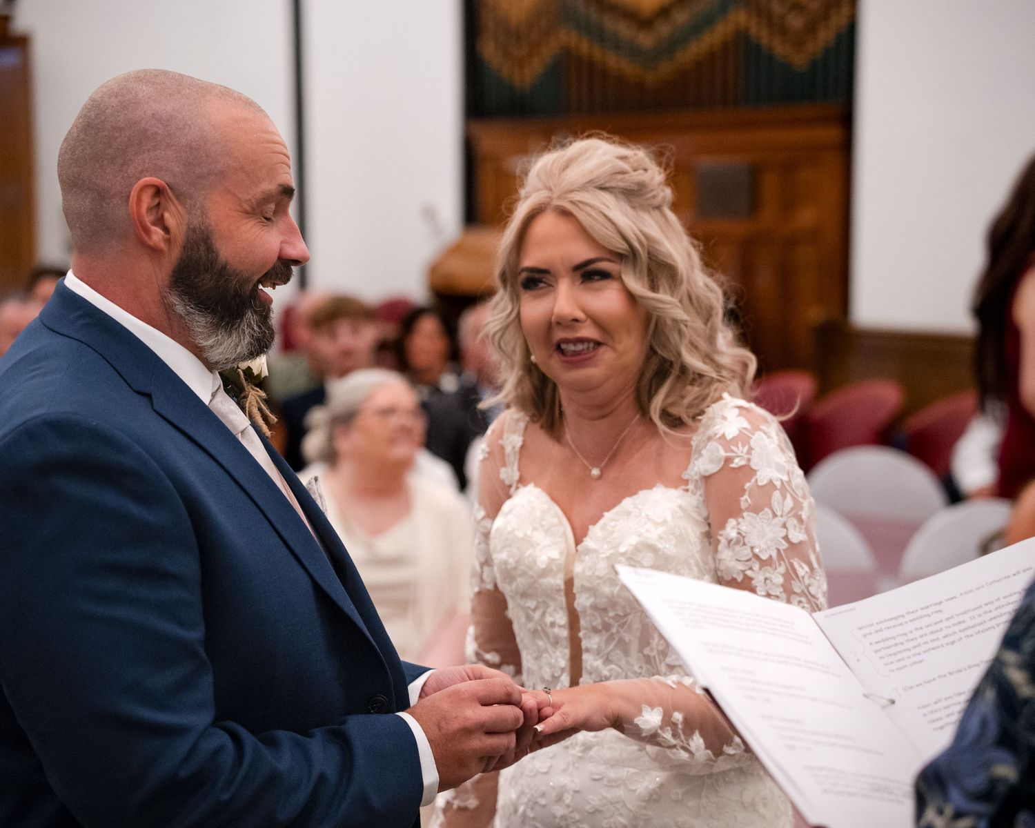 Bride and groom exchange vows during indoor wedding ceremony, bride in lace gown holding papers, groom in navy suit.