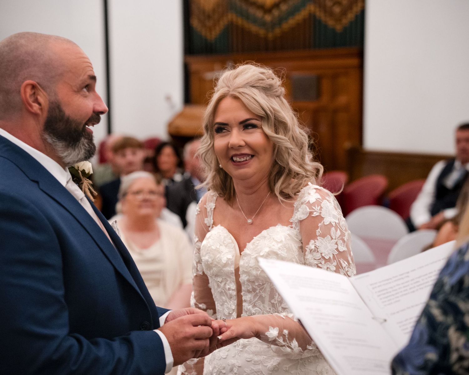 A bride and groom exchange vows at their wedding ceremony, with the smiling bride holding papers in a elegant lace gown.