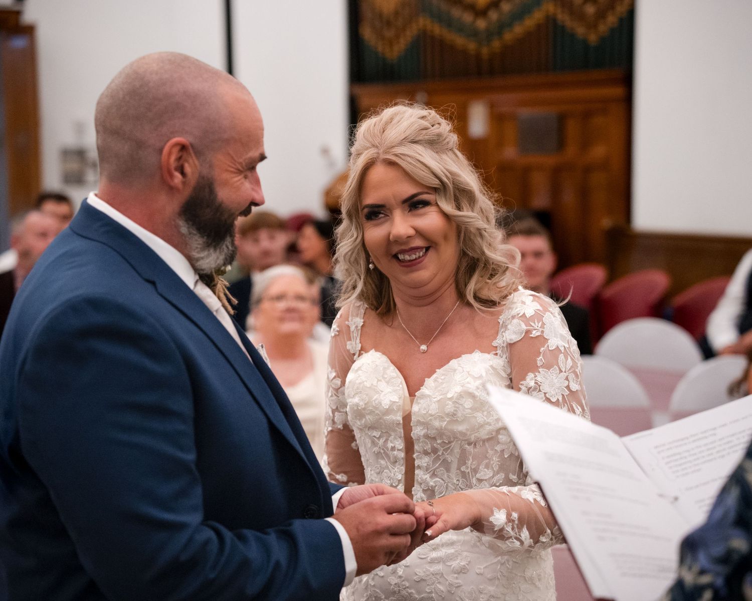 Bride and groom exchanging vows at wedding ceremony, bride wearing lace dress smiling joyfully at altar.