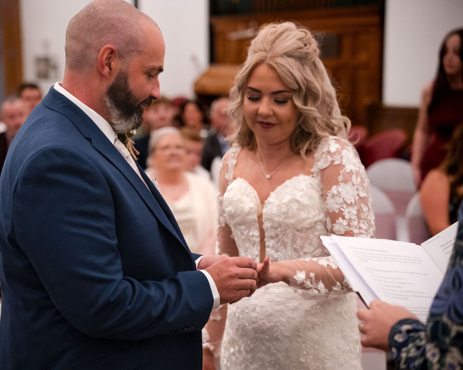 Bride and groom exchanging rings during wedding ceremony, bride in lace gown, groom in navy suit.