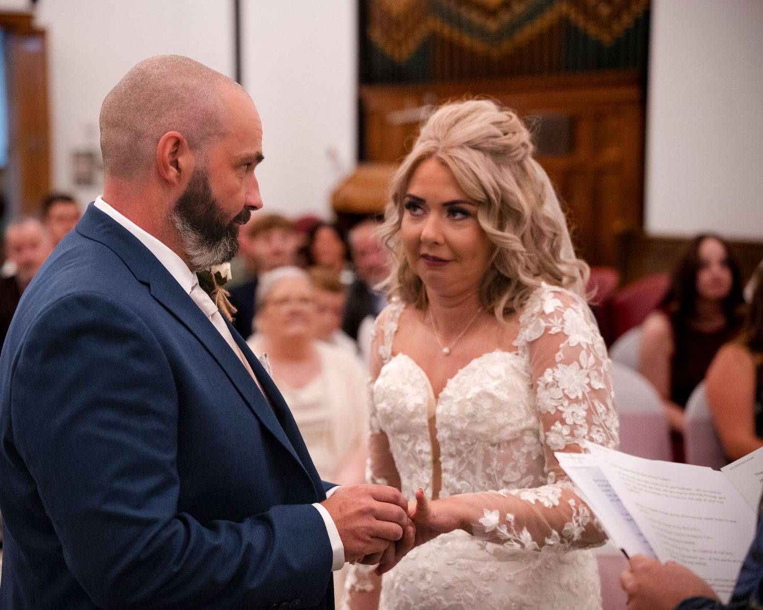 Bride and groom exchanging vows at wedding ceremony, bride in lace dress holding papers, groom in navy suit.