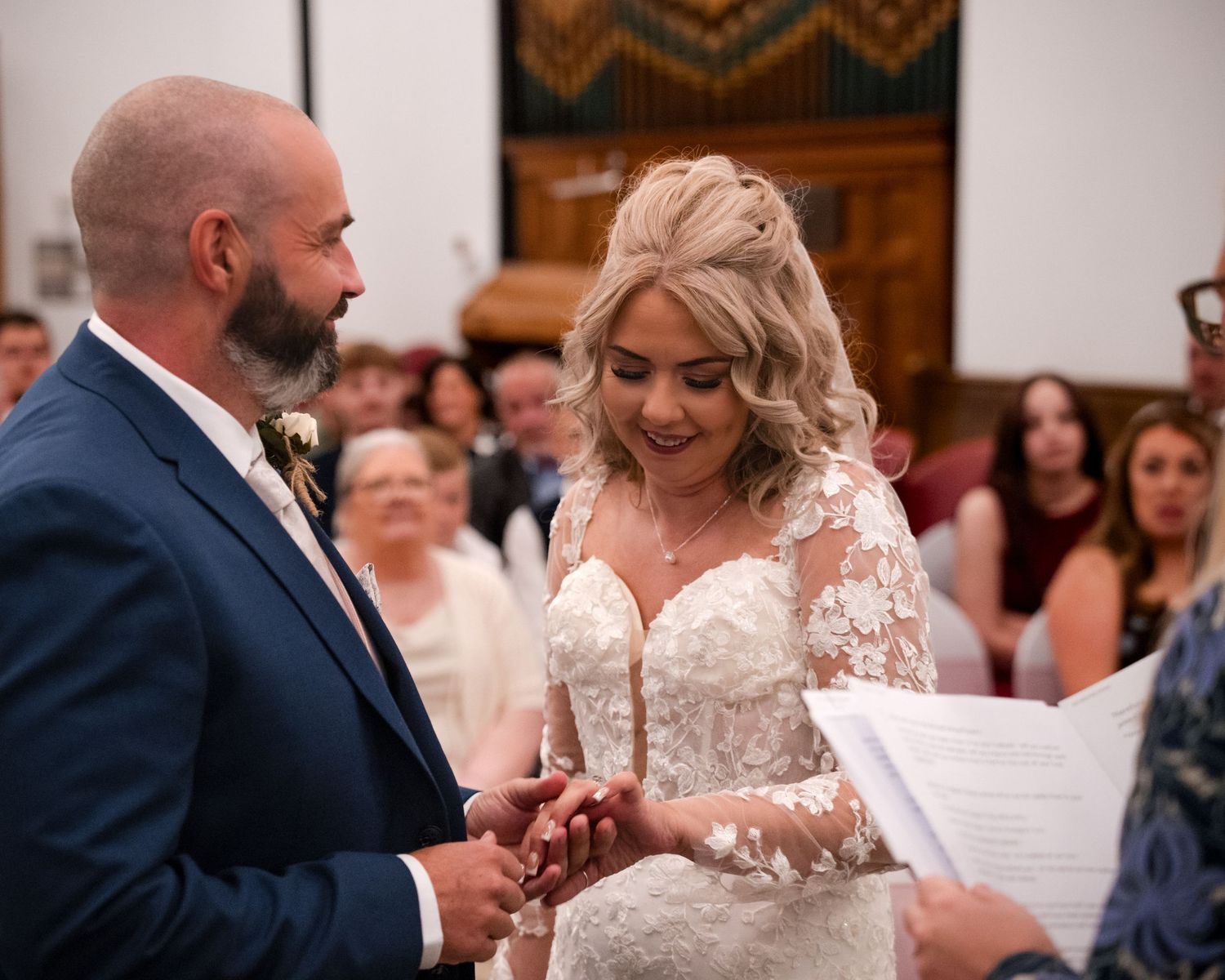 Bride and groom exchanging rings during wedding ceremony in a church, bride wearing lace gown.
