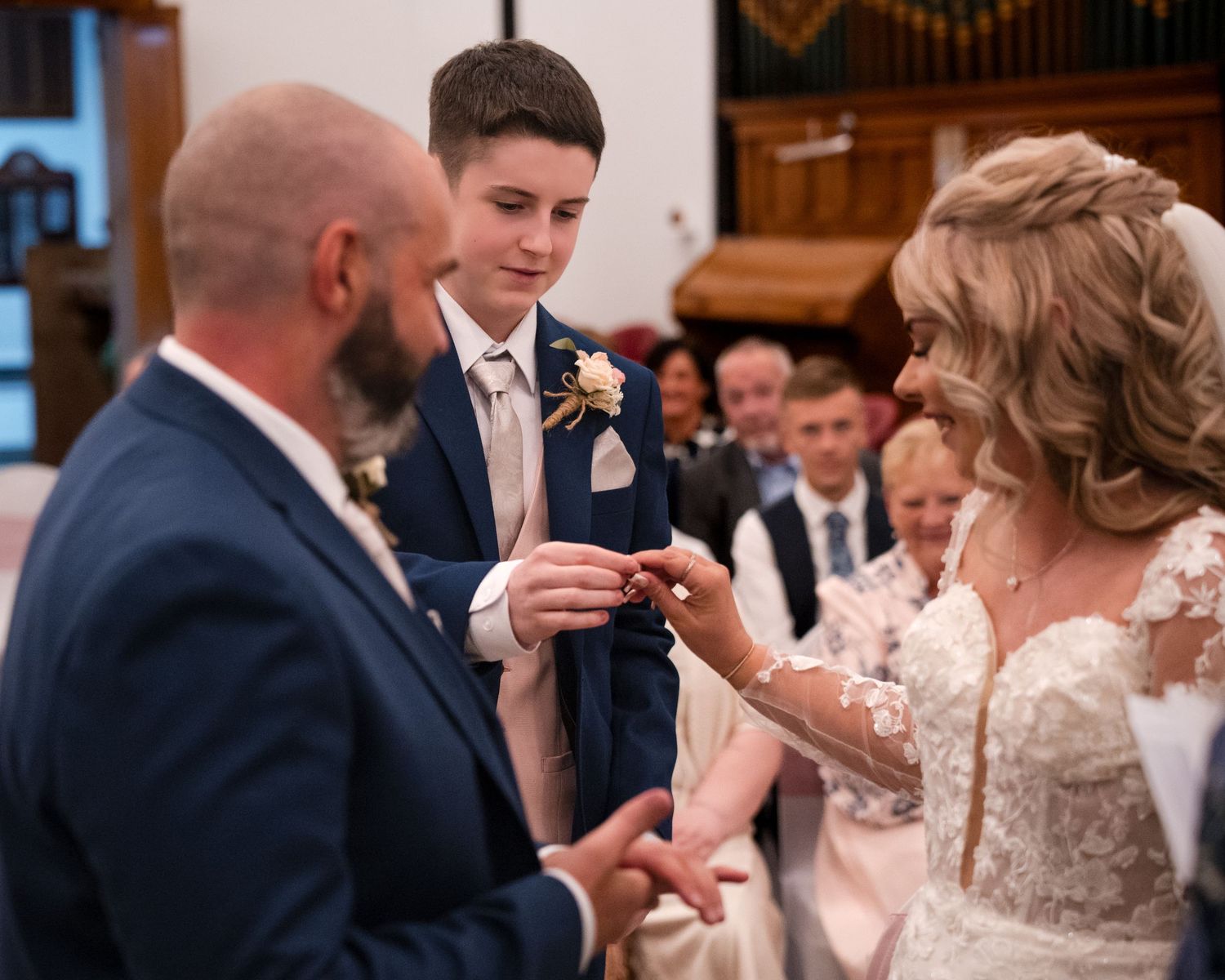 Bride placing wedding ring on groom's finger during church ceremony with young boy holding ring box nearby.