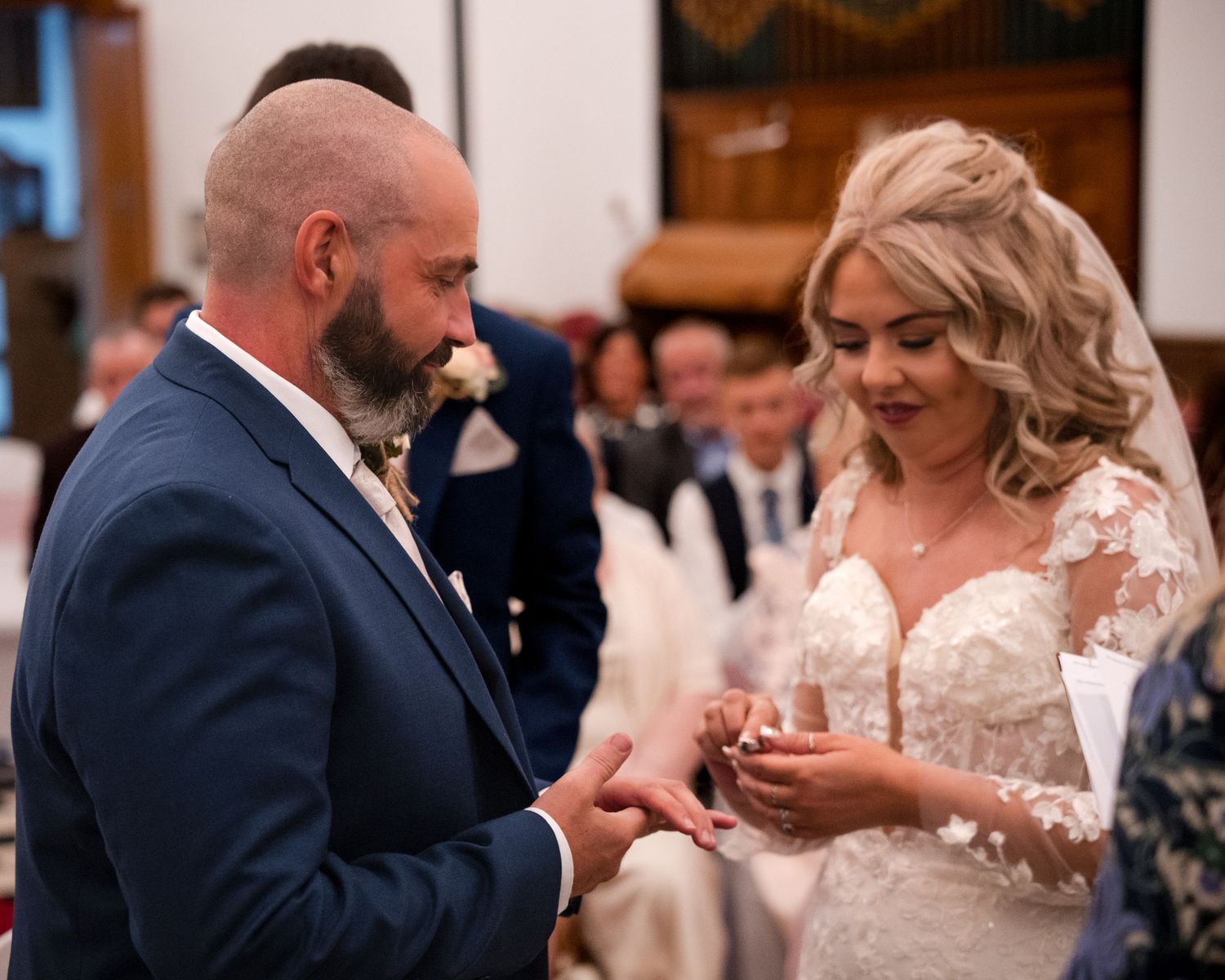 Groom in blue suit placing wedding ring on brides finger during ceremony, bride wearing lace dress.