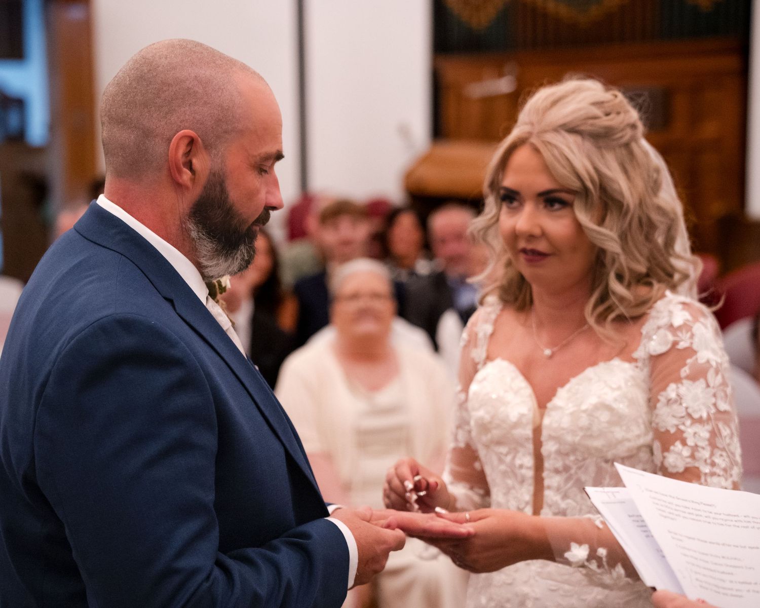 Bride and groom exchanging rings during wedding ceremony in a church, bride wearing lace gown holding paper vows.
