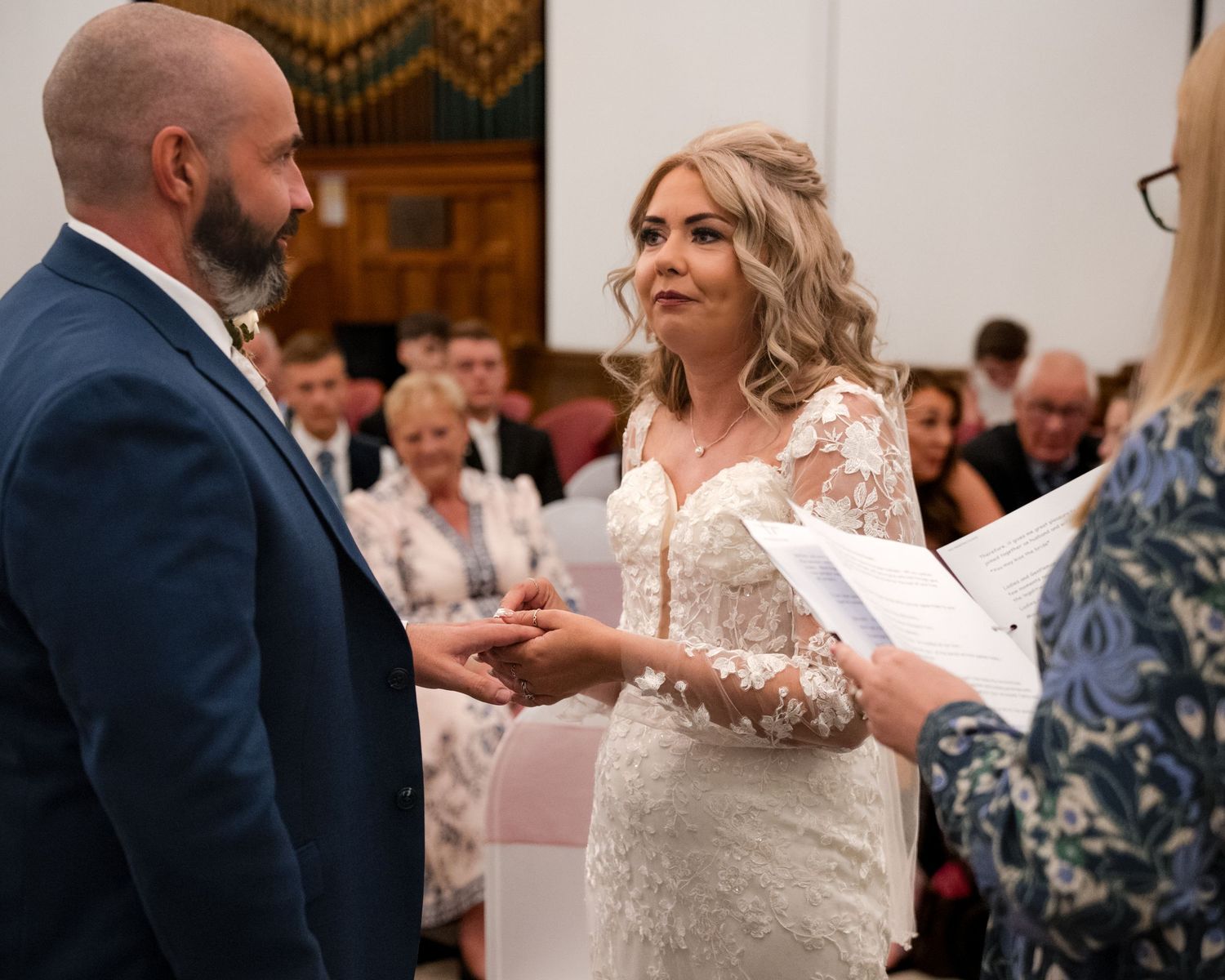 Bride and groom exchange vows during wedding ceremony, bride in lace gown holding hands with groom in navy suit.