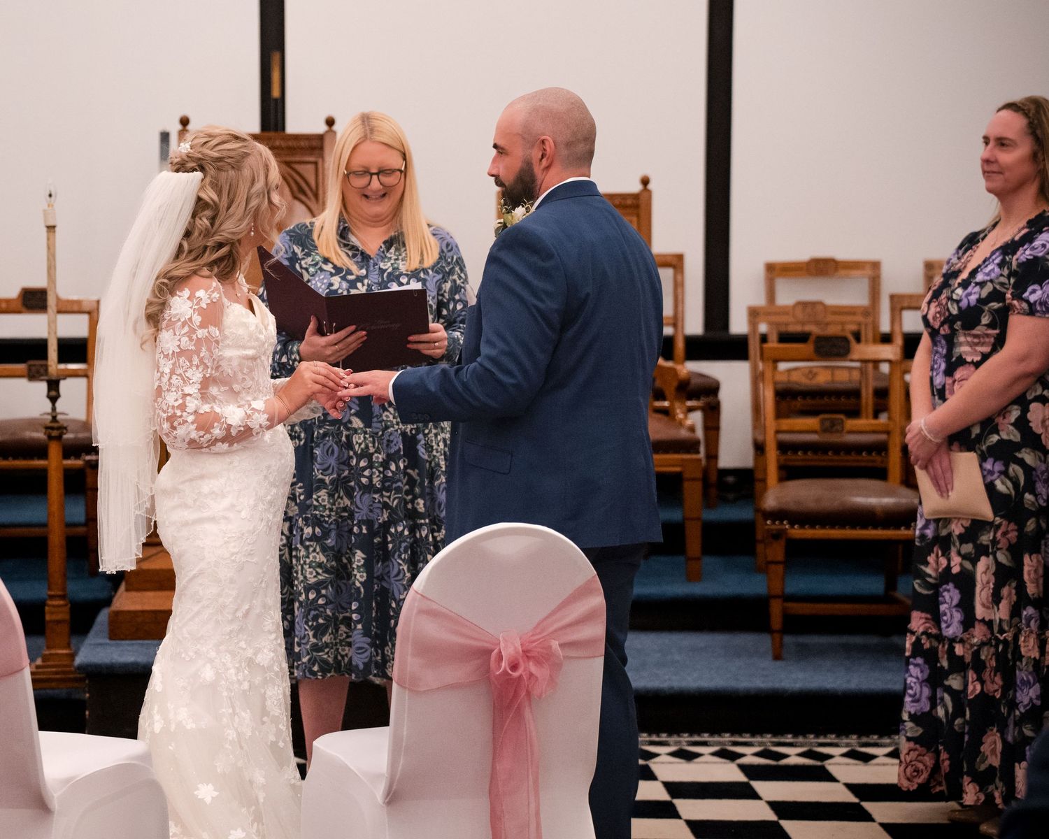 Bride and groom exchanging rings at wedding ceremony with officiant in a chapel.