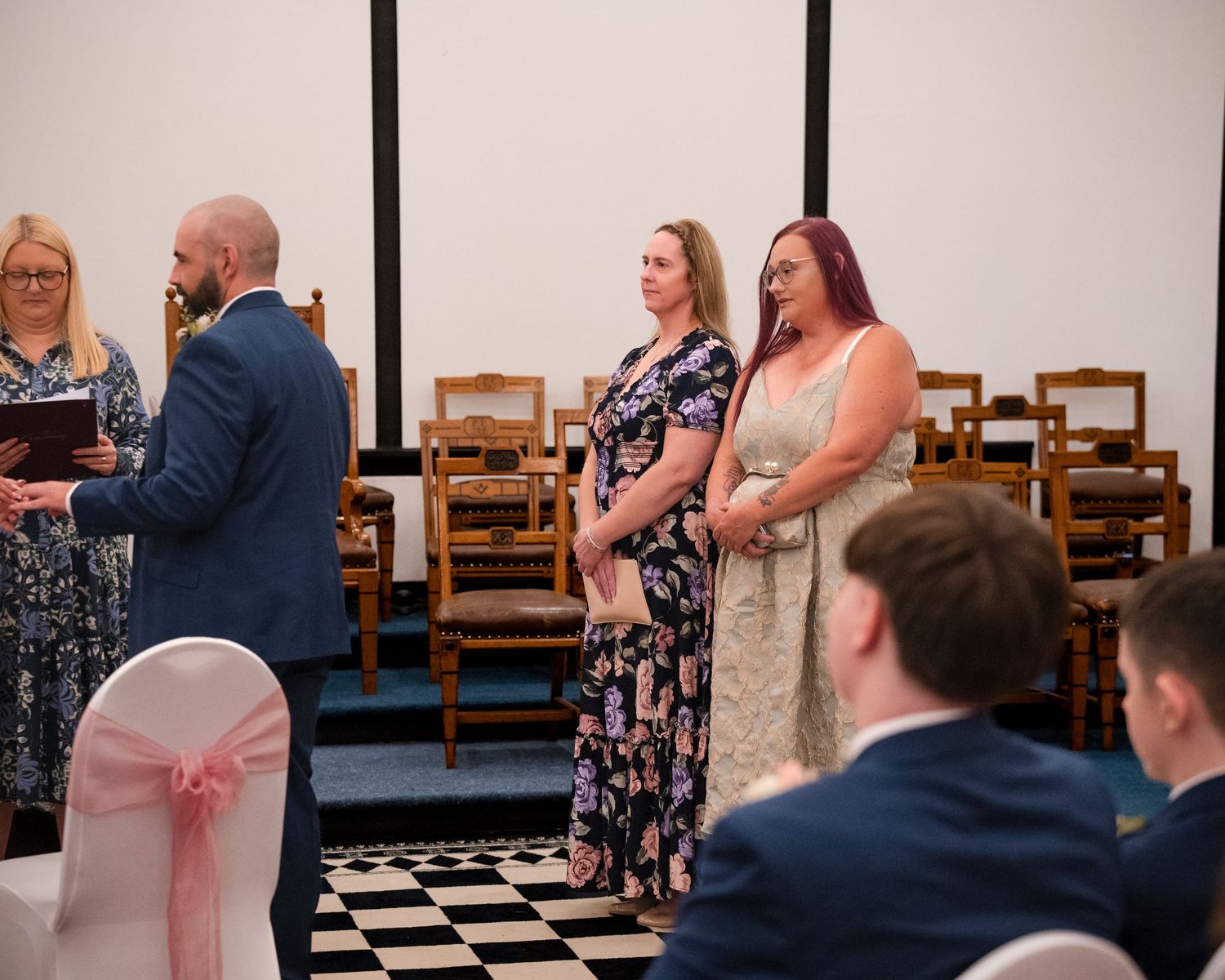 Two women stand at a wedding ceremony in a black and white checkered floor venue with guests seated nearby.