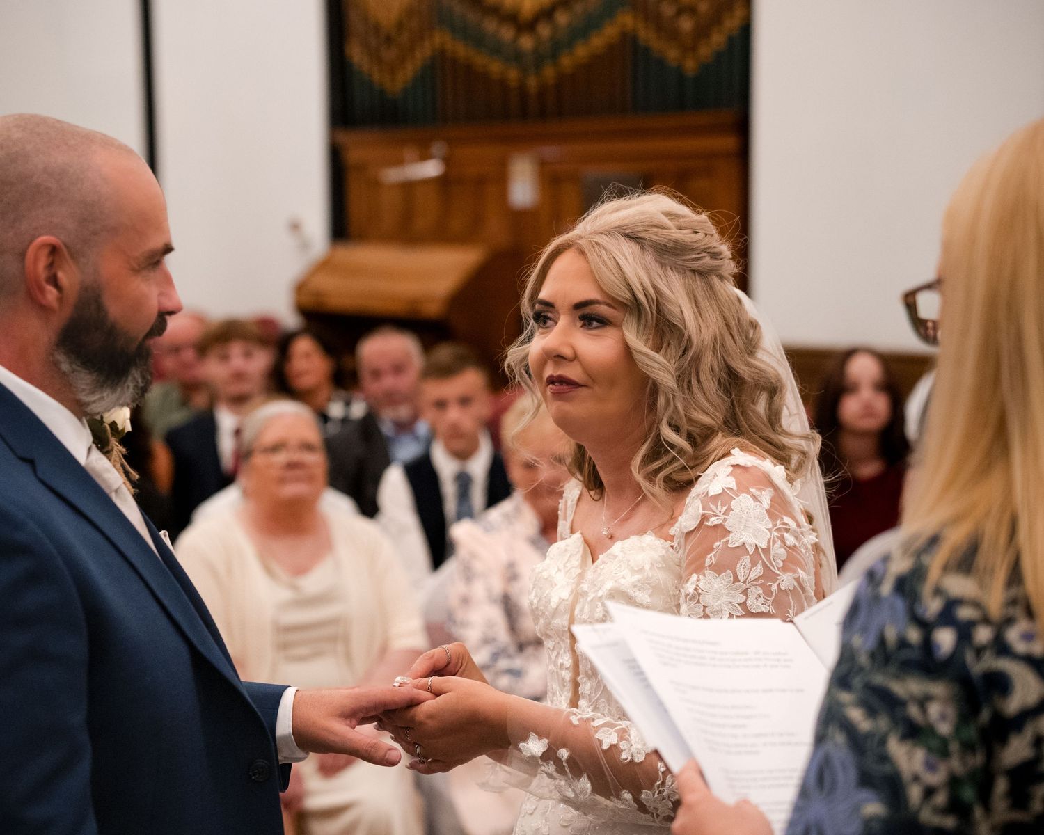 Bride and groom exchange vows during an indoor wedding ceremony, with guests seated in the background.