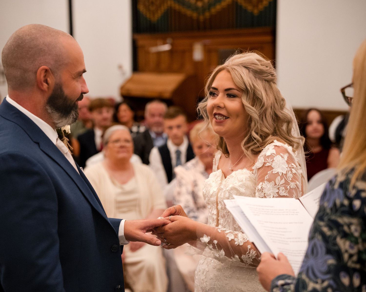 Bride and groom exchanging rings at wedding ceremony in church, bride smiling in lace gown with guests seated behind.