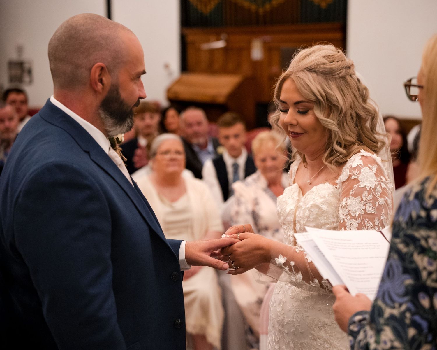 Bride and groom exchanging rings at church wedding ceremony with guests seated in pews behind them.