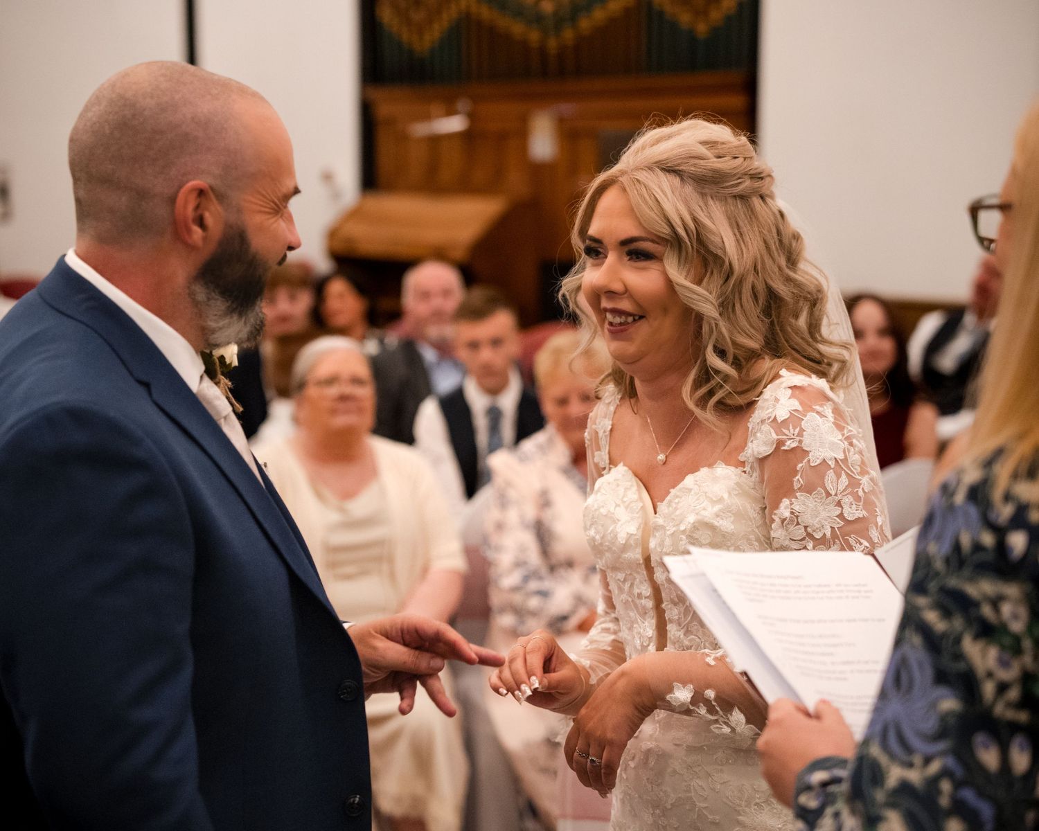 Bride and groom exchange rings at church wedding ceremony, bride smiling in lace gown.
