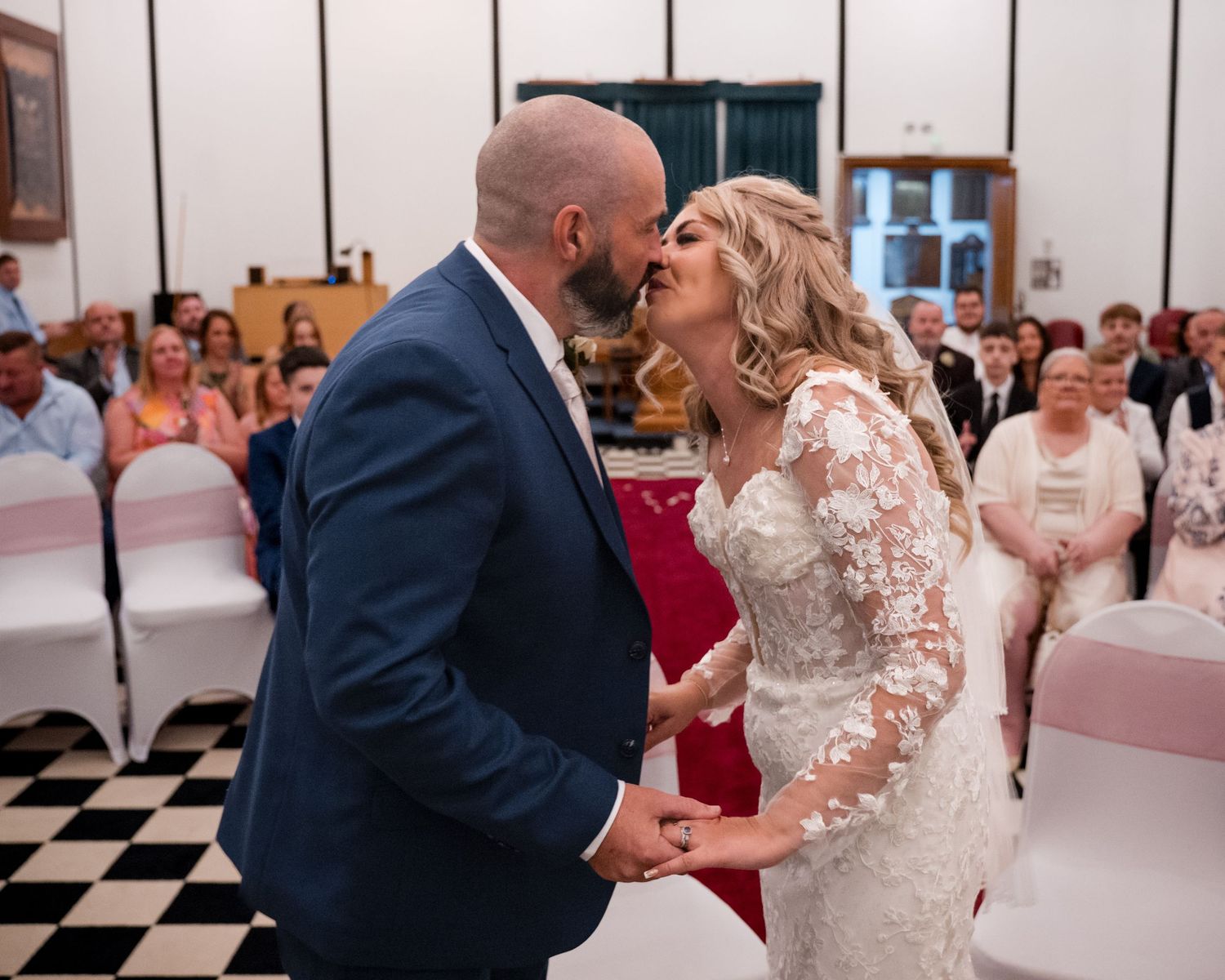 Bride and groom share first kiss at wedding ceremony with guests seated in decorated hall.
