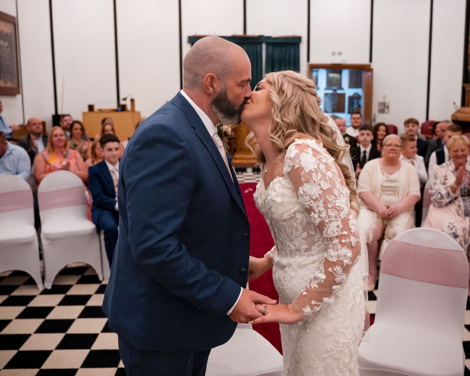 Bride and groom share their first kiss at a wedding ceremony in a hall with checkered floors and seated guests.